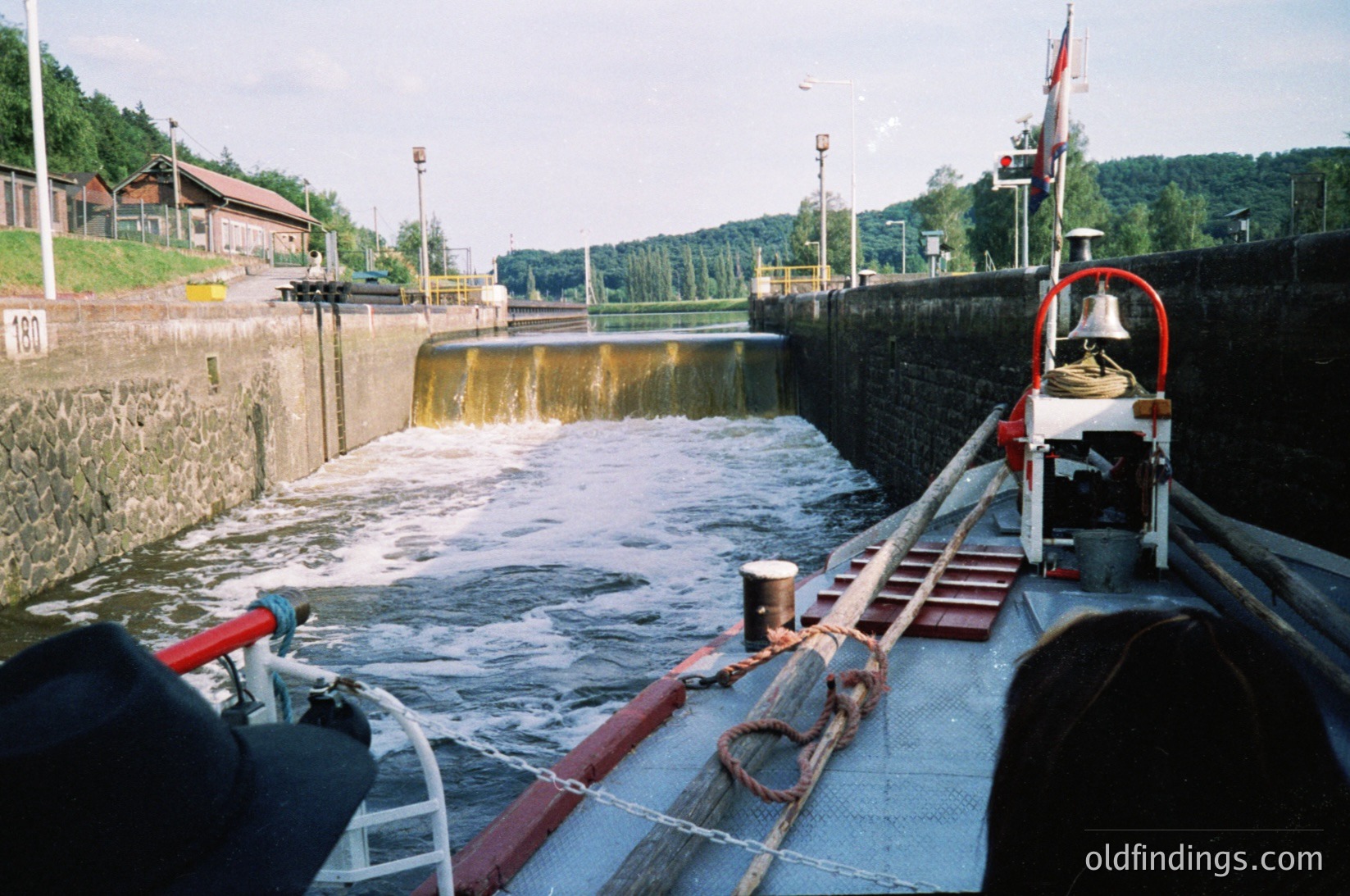 Lock gate in operation with water flow, featuring a red-and-white handrail and bell system. Industrial-era infrastructure with wooden dock and rusted metal fixtures. Surrounding greenery and distant buildings suggest a rural or small-town setting.