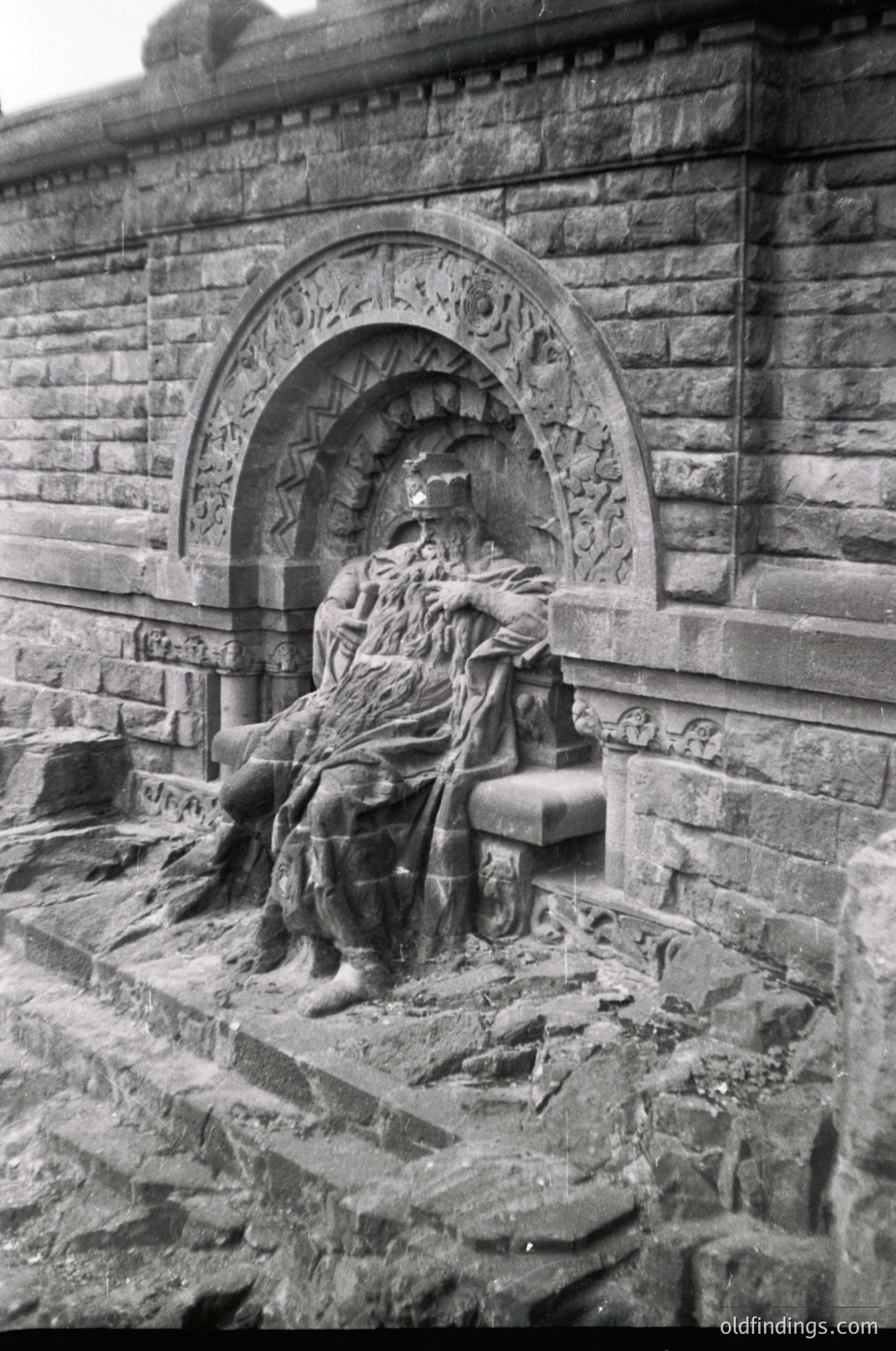 Monolithic stone relief sculpture of a seated figure draped in flowing robes, integrated into a stone archway with intricate carvings. Likely a memorial or commemorative monument, featuring classical motifs.