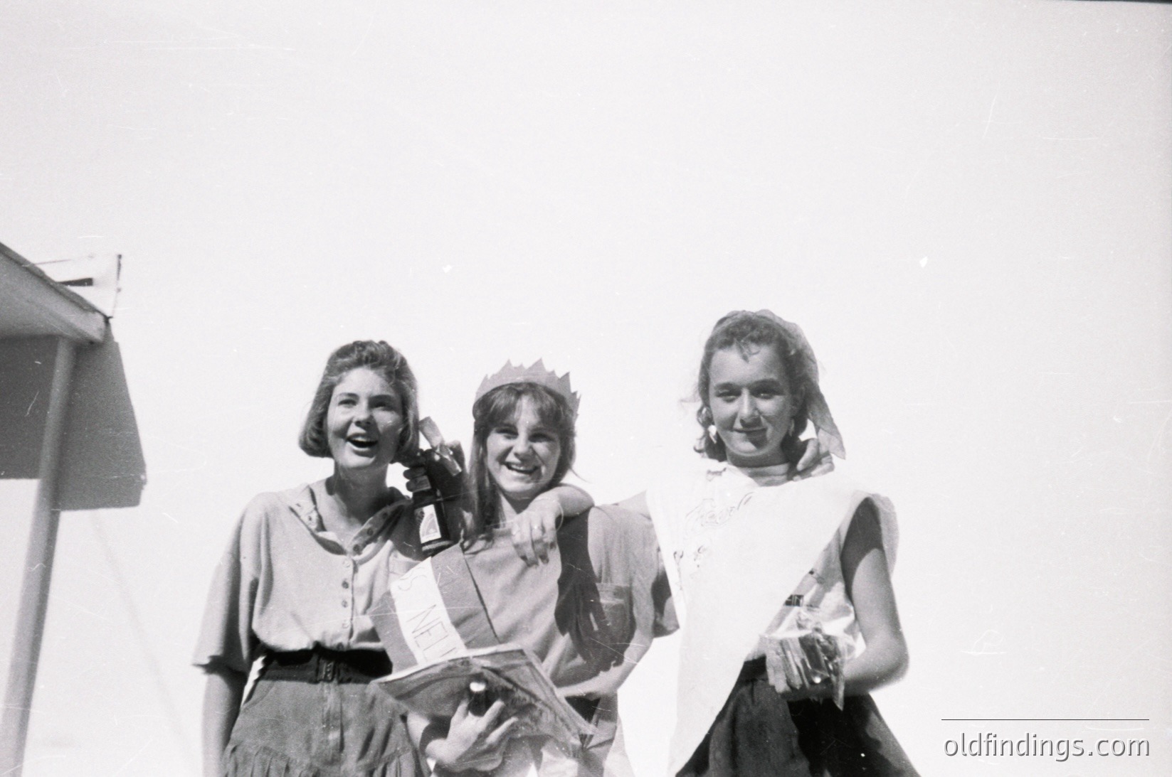 Three women pose outdoors in mid-20th century attire, likely 1950s–1960s. Center figure wears a paper crown and sash, suggesting a pageant or celebration. Left wears a blouse with a belted waist, right a sheer veil over a dress. Background shows a plain wall and partial structure, indicating an event space.