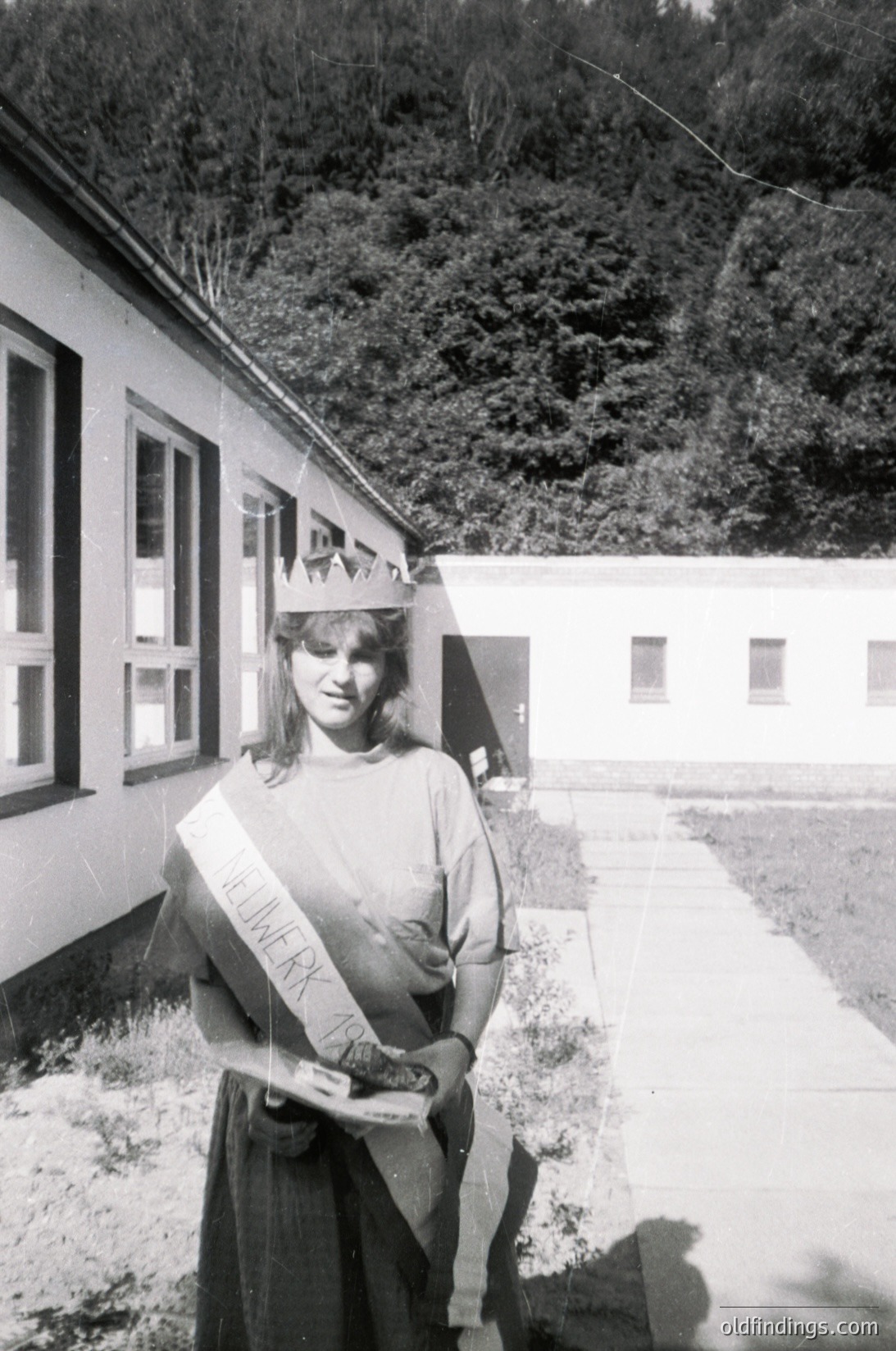Young woman in 1970s-era sash and crown, likely a beauty pageant contestant, posing outdoors near a plain, institutional-style building. Text on sash reads "Miss Winter." Forested hillside in background suggests a rural or suburban setting.