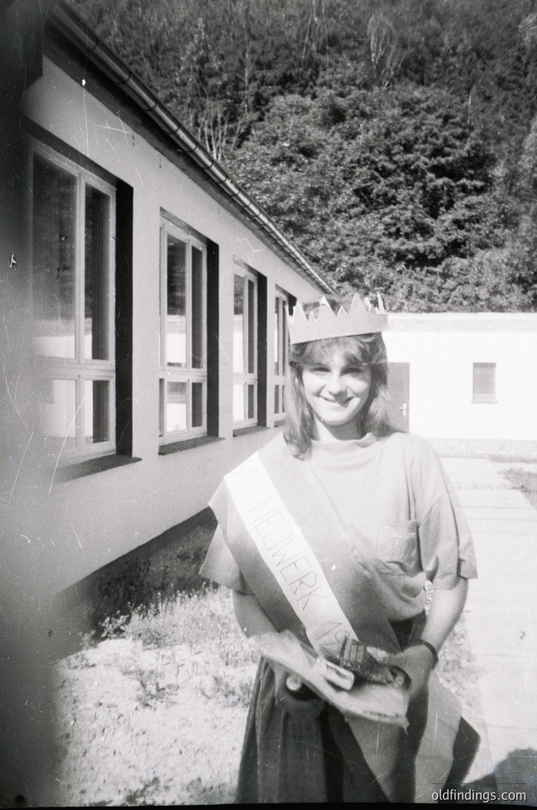Young woman in graduation regalia poses outside a utilitarian building, likely a school or dormitory, with forested hills in background. Mid-20th century academic attire includes a sash and crown, suggesting a formal ceremony.