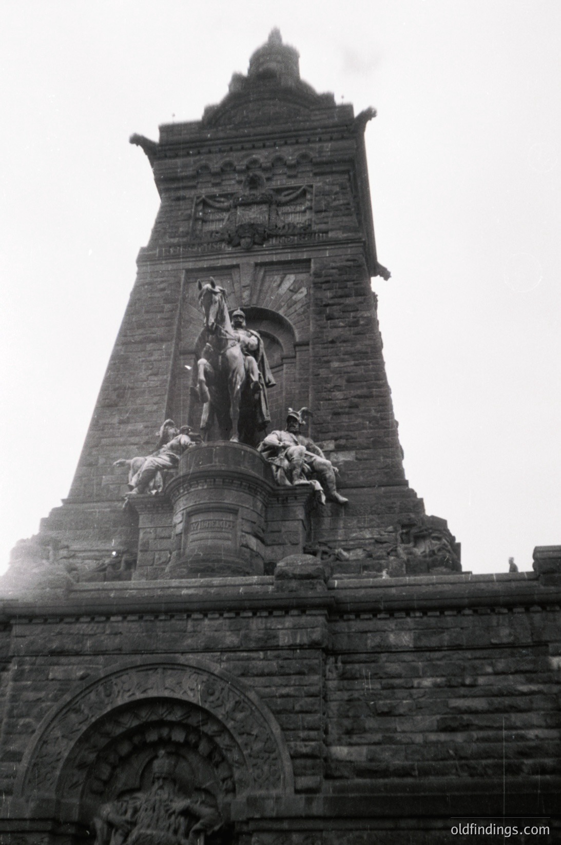 Monumental equestrian statue atop a stone tower, likely 19th-century European. Central figure on horseback flanked by kneeling soldiers, set against a Gothic-inspired facade. Intricate carvings and weathered stone suggest historical significance.