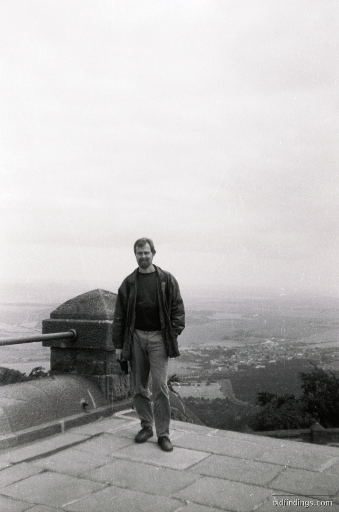 Black-and-white portrait of a man in 1970s-era attire (leather jacket, rolled-up trousers) standing atop a stone terrace with panoramic cityscape below. Overcast skies and misty horizon suggest early morning or late afternoon. Urban sprawl hints at European cityscape, possibly Eastern Bloc era.