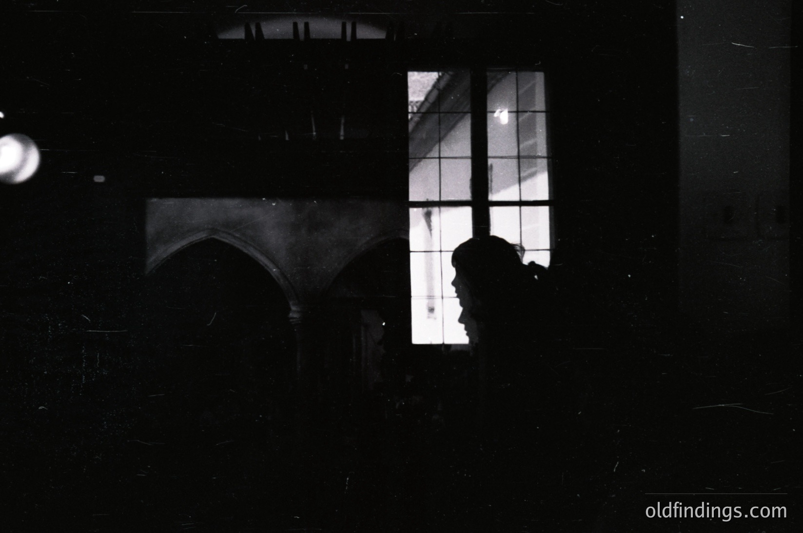 Silhouette of a person framed by a large arched window with grid-patterned glass, reflecting faint architectural details. High-contrast black-and-white composition evokes mid-20th century urban photography.