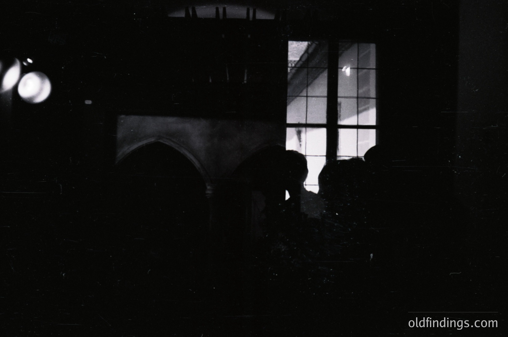 Silhouette of a person standing near a window with grid-patterned glass, illuminated from behind by artificial light. Reflections of architectural elements and light fixtures create abstract shapes on the wall. High-contrast black-and-white composition suggests a 20th-century urban setting, possibly a subway or train station. &W
