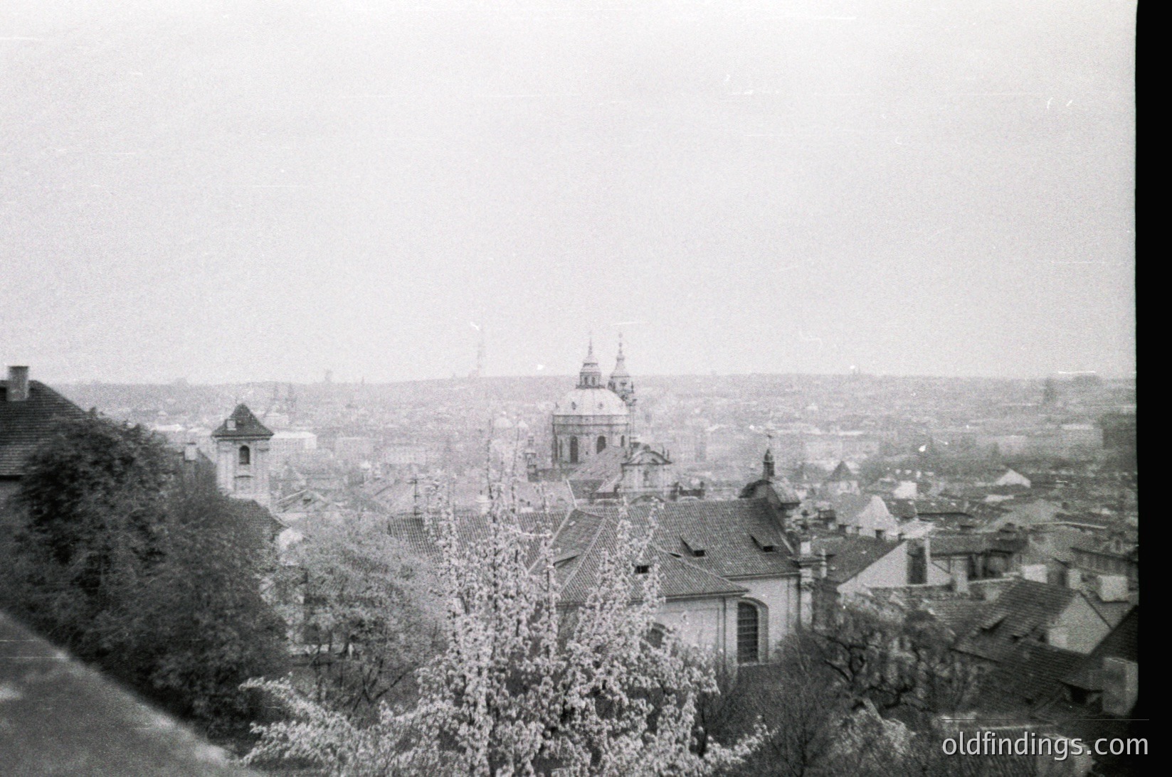 Early 20th-century urban panorama featuring a domed church with cross atop a hill, surrounded by low-rise buildings with pitched roofs. Dense tree line in foreground suggests elevated vantage point. Foggy atmosphere enhances historical ambiance.