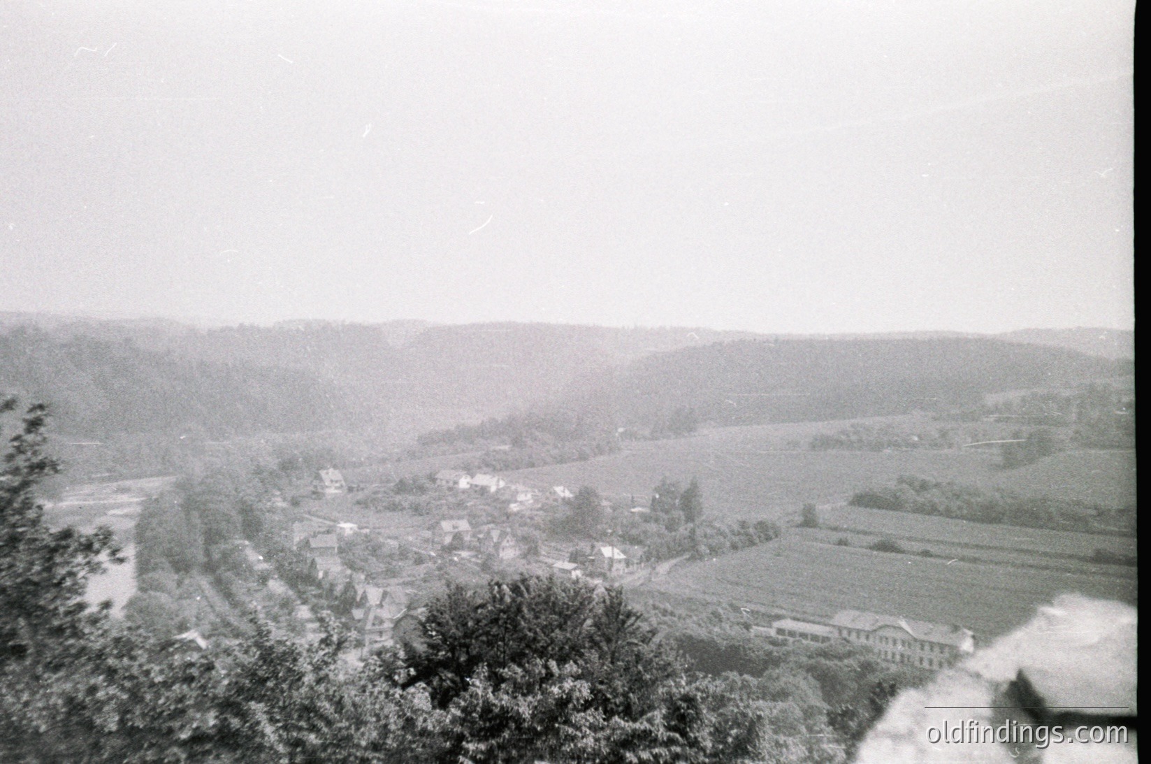 Vintage black-and-white aerial view of a rural valley with winding roads, clustered buildings, and agricultural fields. Dense forest lines the ridges, framing the scene. Likely early-to-mid 20th century European countryside.