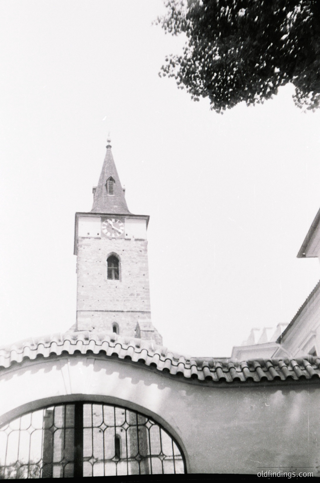 Mid-century church tower with pointed steeple and arched bell window, framed by ornate wrought-iron gate. Stone façade and tiled roof suggest Eastern European architecture. Likely –1960s era.