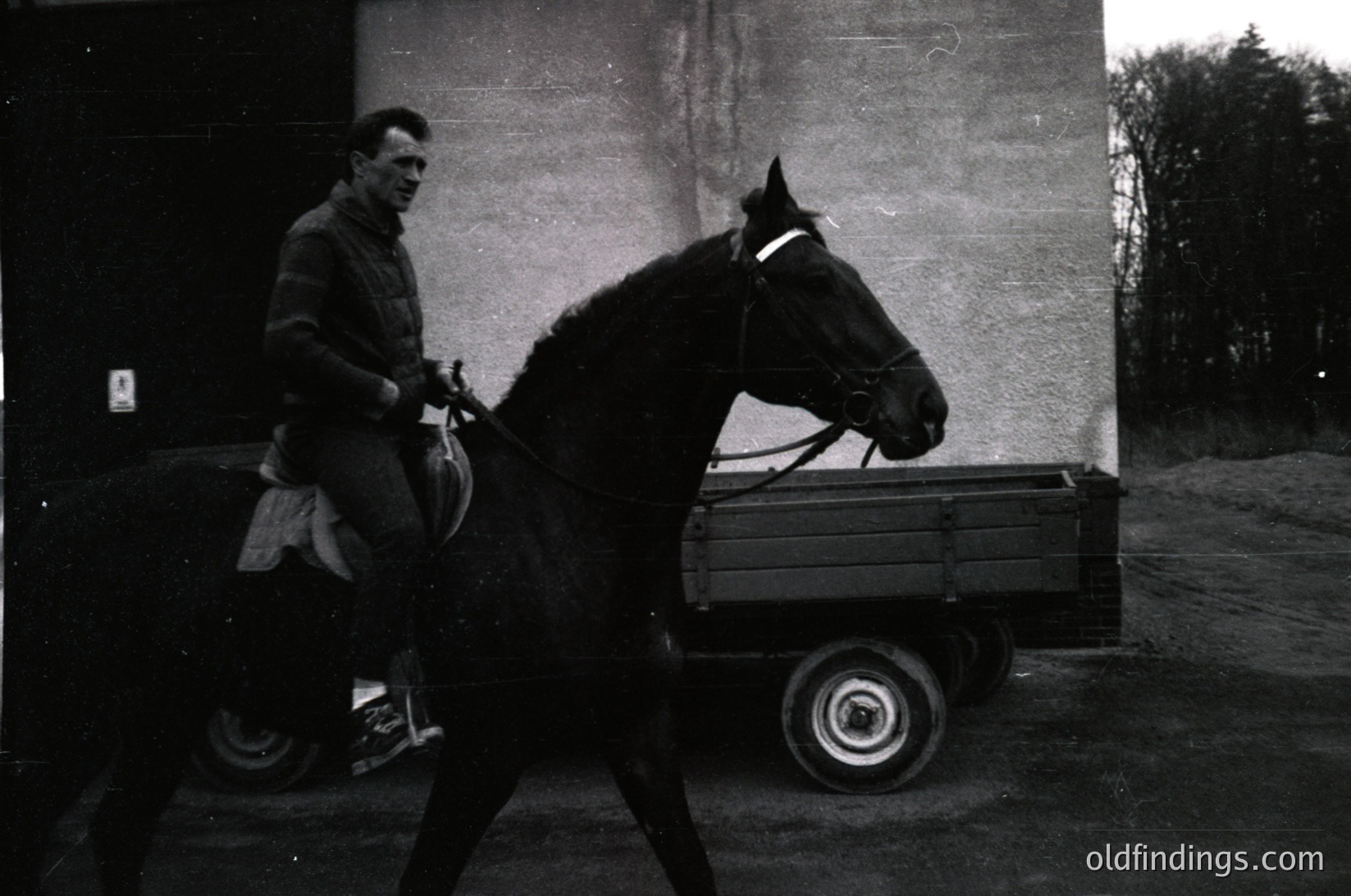 Man in 1950s-60s workwear leads a draft horse pulling a wooden cart with large spoked wheels. Industrial brick wall and rural roadside setting suggest agricultural labor.
