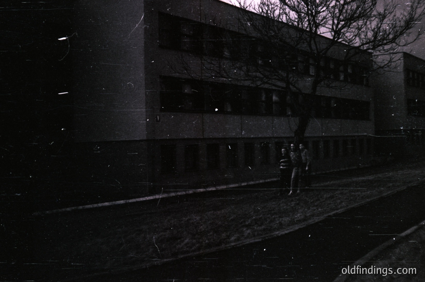Mid-century brutalist concrete building with vertical window bands, captured in monochrome. Two figures in dark coats walk along a wet pavement under bare trees, suggesting winter or early evening. Urban architecture with stark geometric lines and minimalist design.
