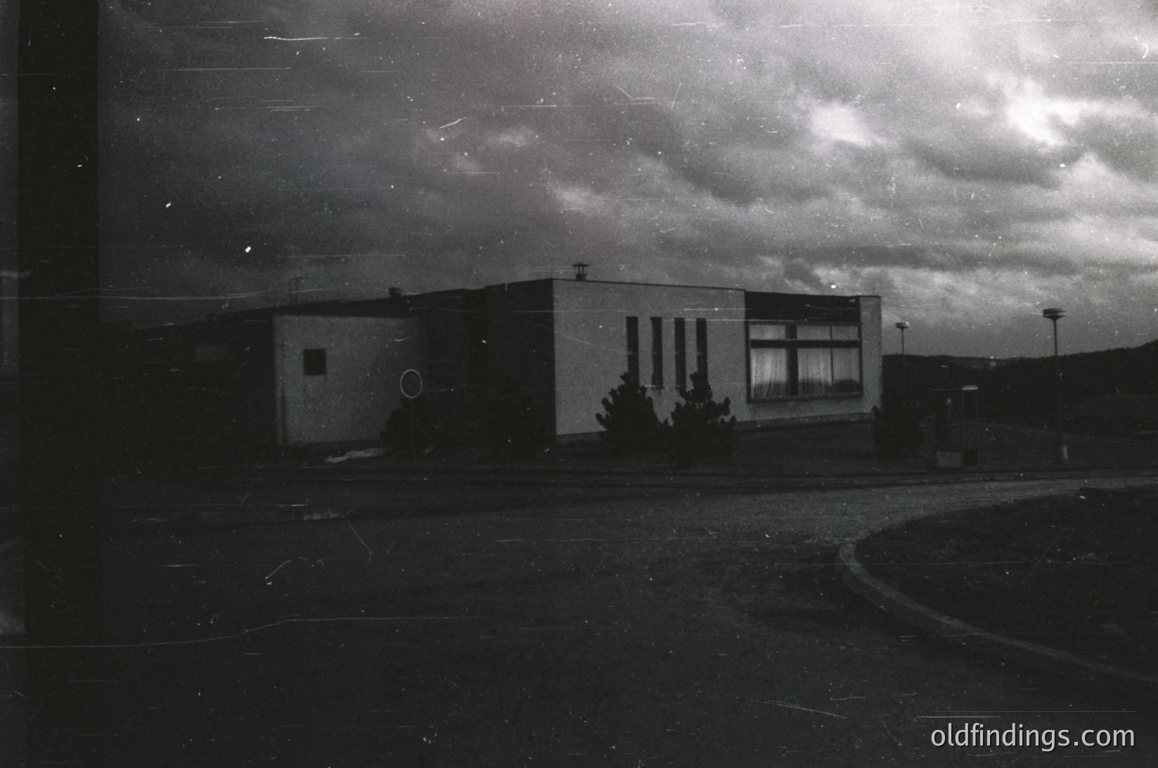 Vintage black-and-white photo of a low-rise, rectangular building with minimalist Brutalist concrete façade and circular window. Overcast sky and empty paved area suggest mid-20th century urban/suburban design.