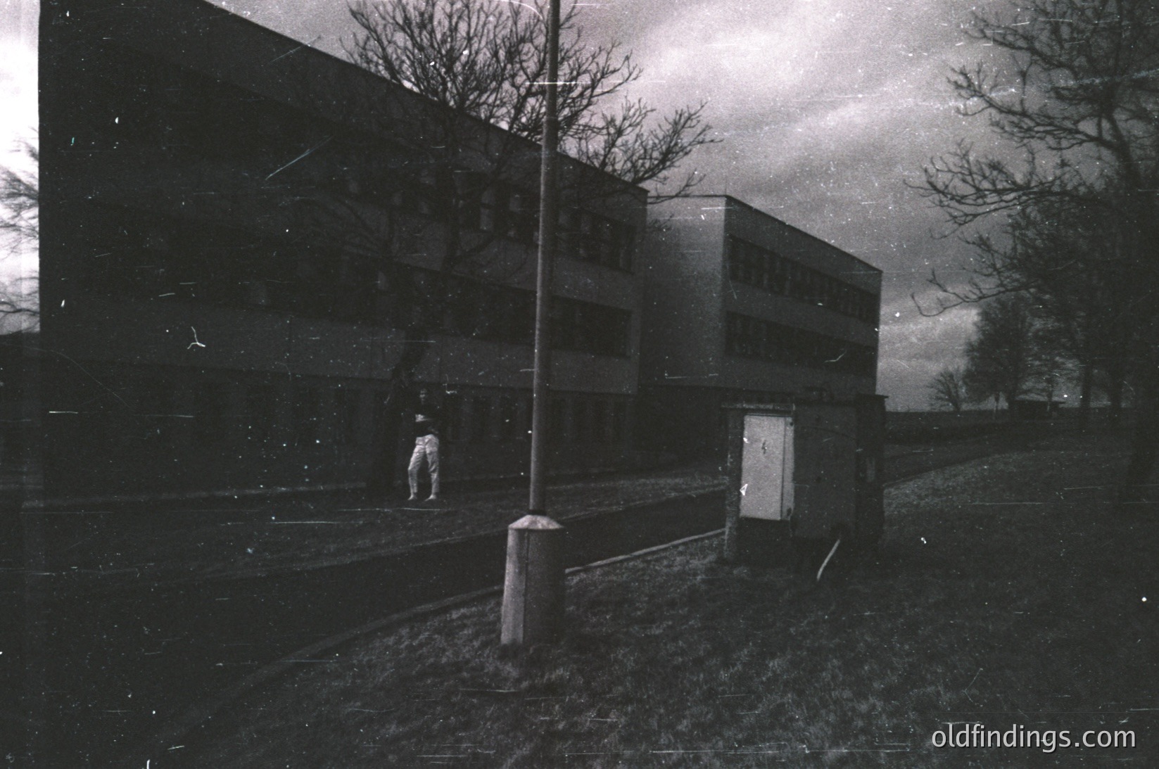 Mid-century brutalist apartment block in monochrome, featuring raw concrete surfaces and geometric lines. A lone figure in light clothing walks toward the building, framed by a utility pole and open gate. Overcast sky enhances the stark, industrial atmosphere. Likely Eastern European, 1960s–1970s.