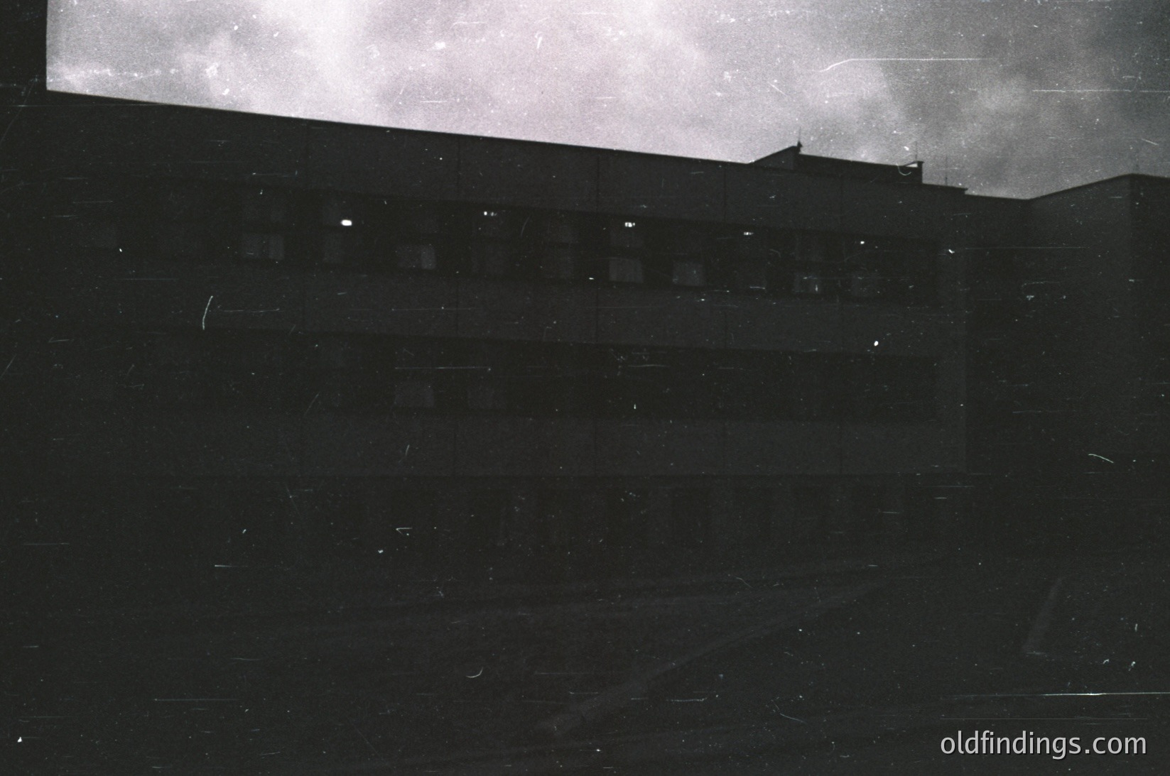 High-contrast black-and-white shot of a multi-story building’s rooftop edge, likely from a low-angle perspective. Horizontal lines emphasize architectural repetition; faint light reveals rooftop details and distant horizon.