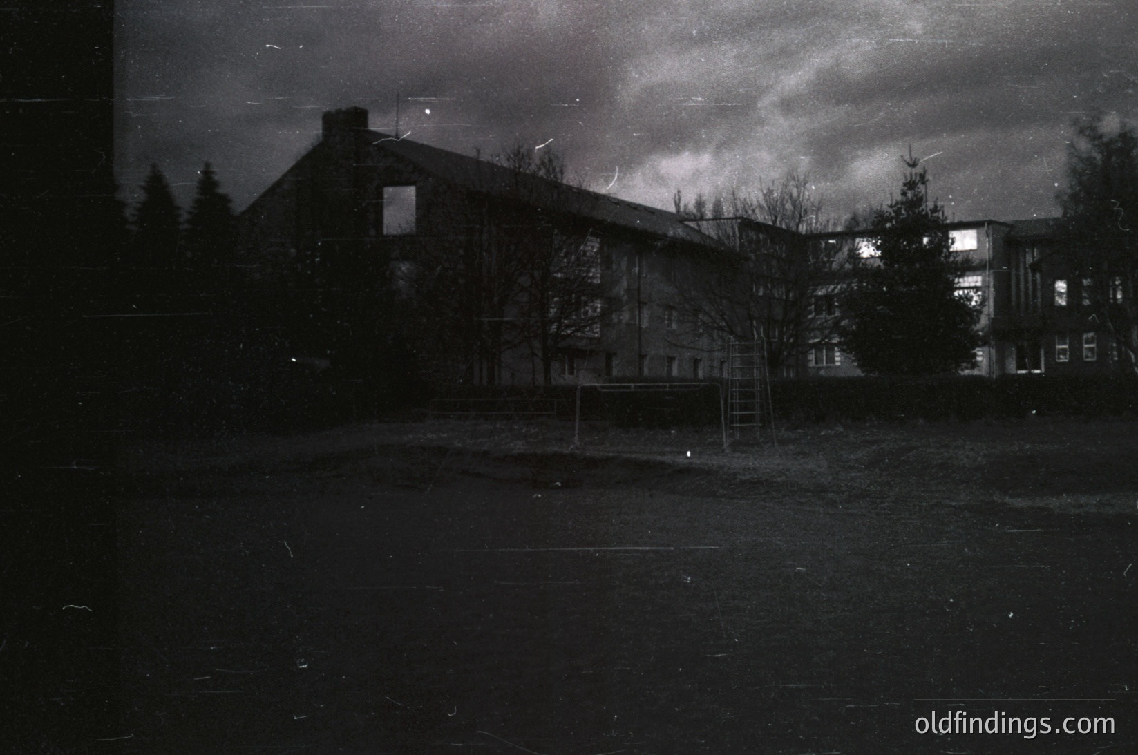 Mid-century concrete apartment block with minimalist Brutalist design, featuring exposed concrete walls and a flat roof. Bare trees and a small sports area suggest winter or early spring. Dramatic lighting and grainy texture indicate vintage film or scanning. Likely Eastern Bloc-era residential architecture.
