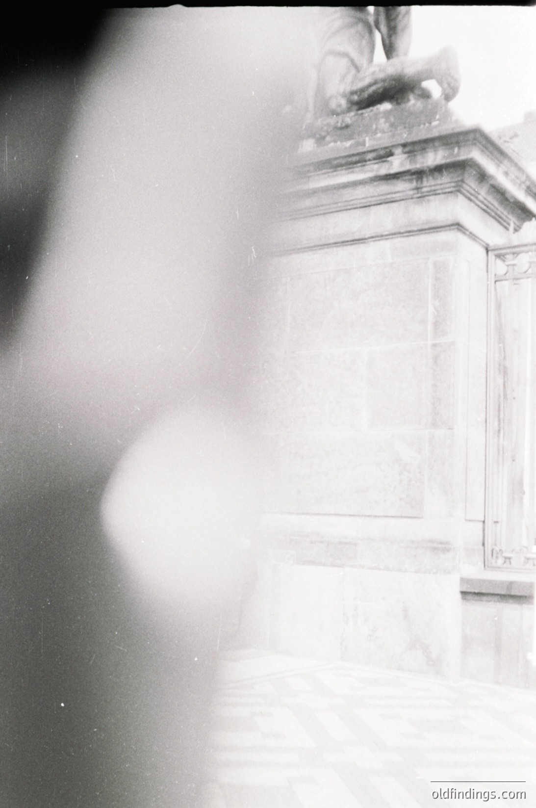 Blurred black-and-white close-up of a classical stone monument featuring a seated figure with wings, likely a cherub or angel. Engraved text on the pedestal suggests commemorative or memorial intent. Art deco-inspired details hint at early-to-mid 20th century craftsmanship.