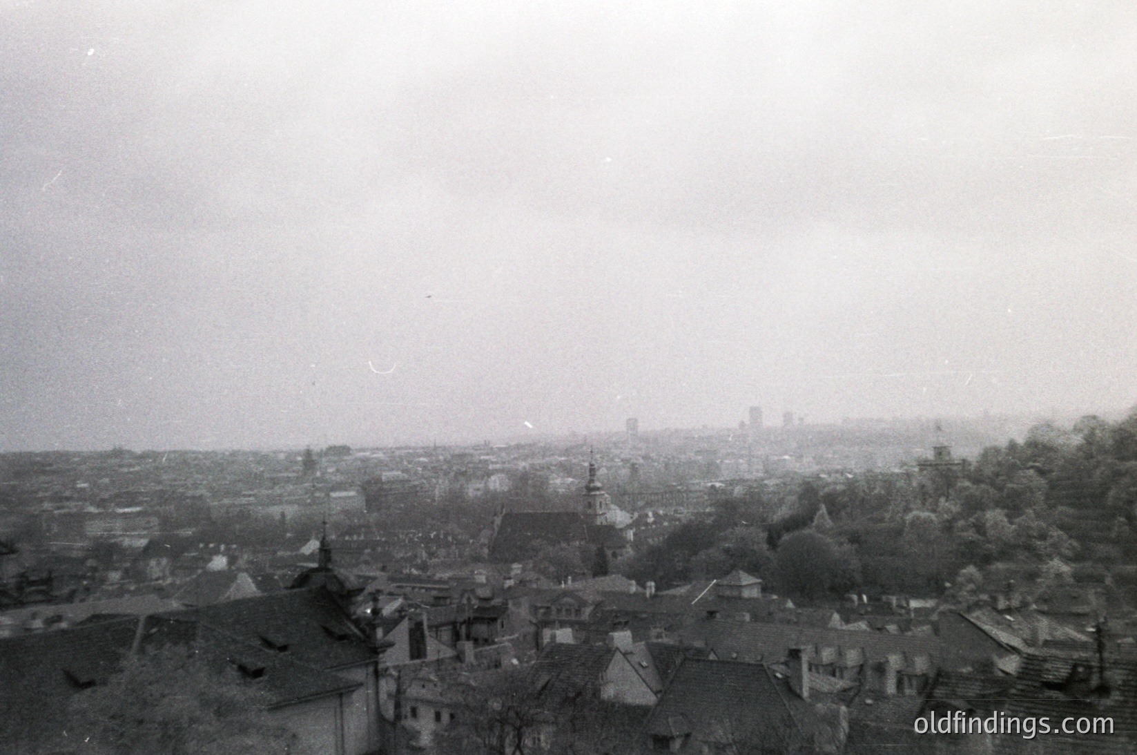 Vintage aerial view of a European cityscape with dense, low-rise architecture and a prominent church spire. Fog obscures distant skyline, emphasizing urban density. Likely mid-20th century due to architectural style and monochrome quality.
