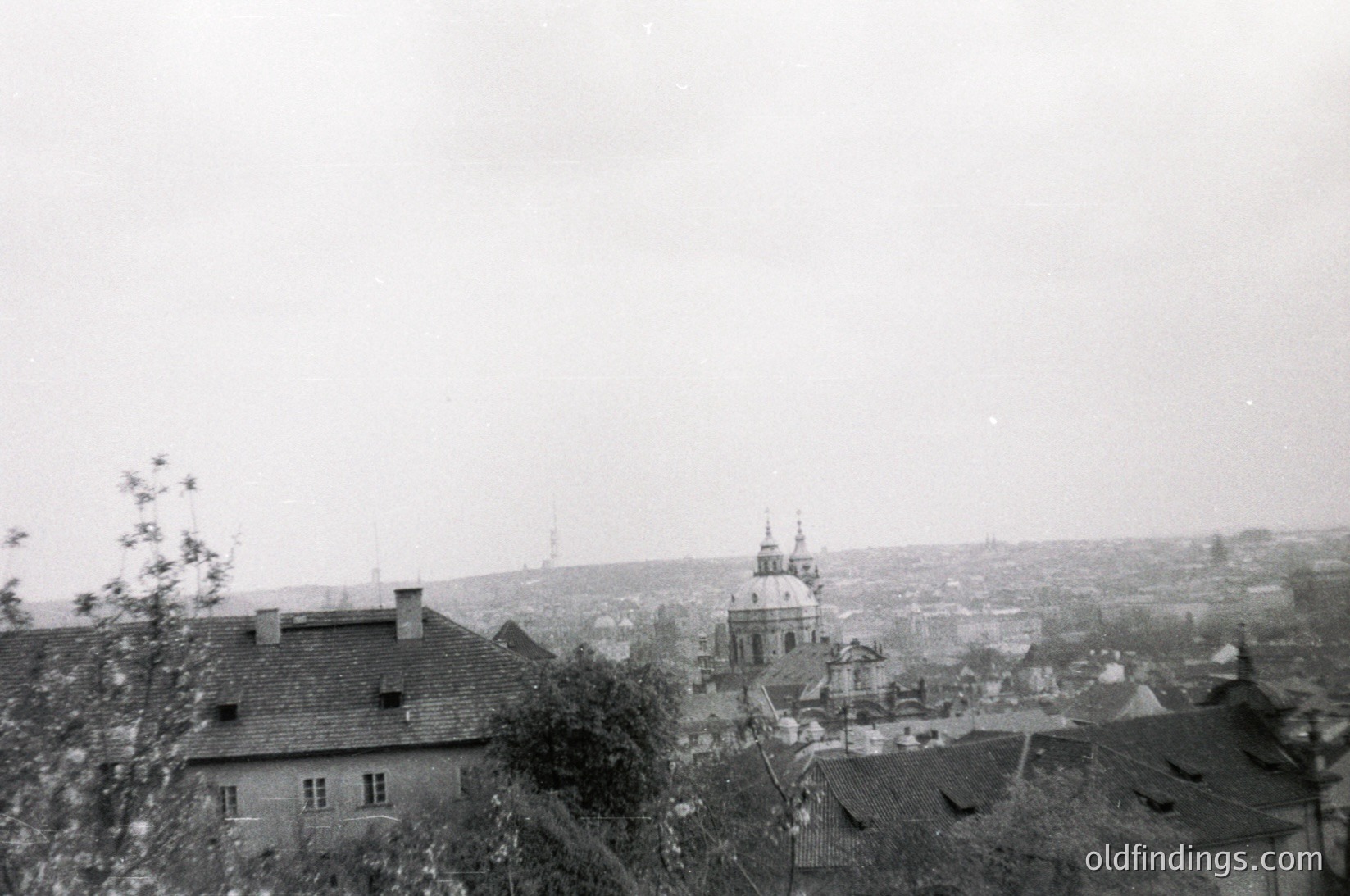 Vintage black-and-white urban landscape featuring a domed church with a tall spire in the background, surrounded by low-rise brick buildings with sloped roofs. Dense foliage partially obscures foreground structures. Overcast sky enhances muted tones. Likely Eastern European setting, mid-20th century.