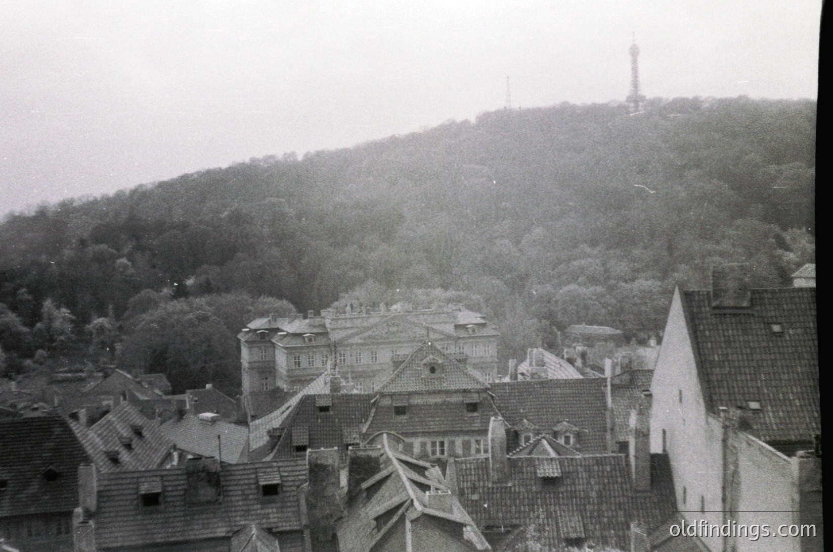 Vintage black-and-white aerial view of a European hillside town with dense, sloped rooftops and a prominent hilltop tower. Architectural style suggests 19th-century urban planning. Fog obscures distant hills, enhancing atmospheric depth. Potential research value for historical urban development studies.
