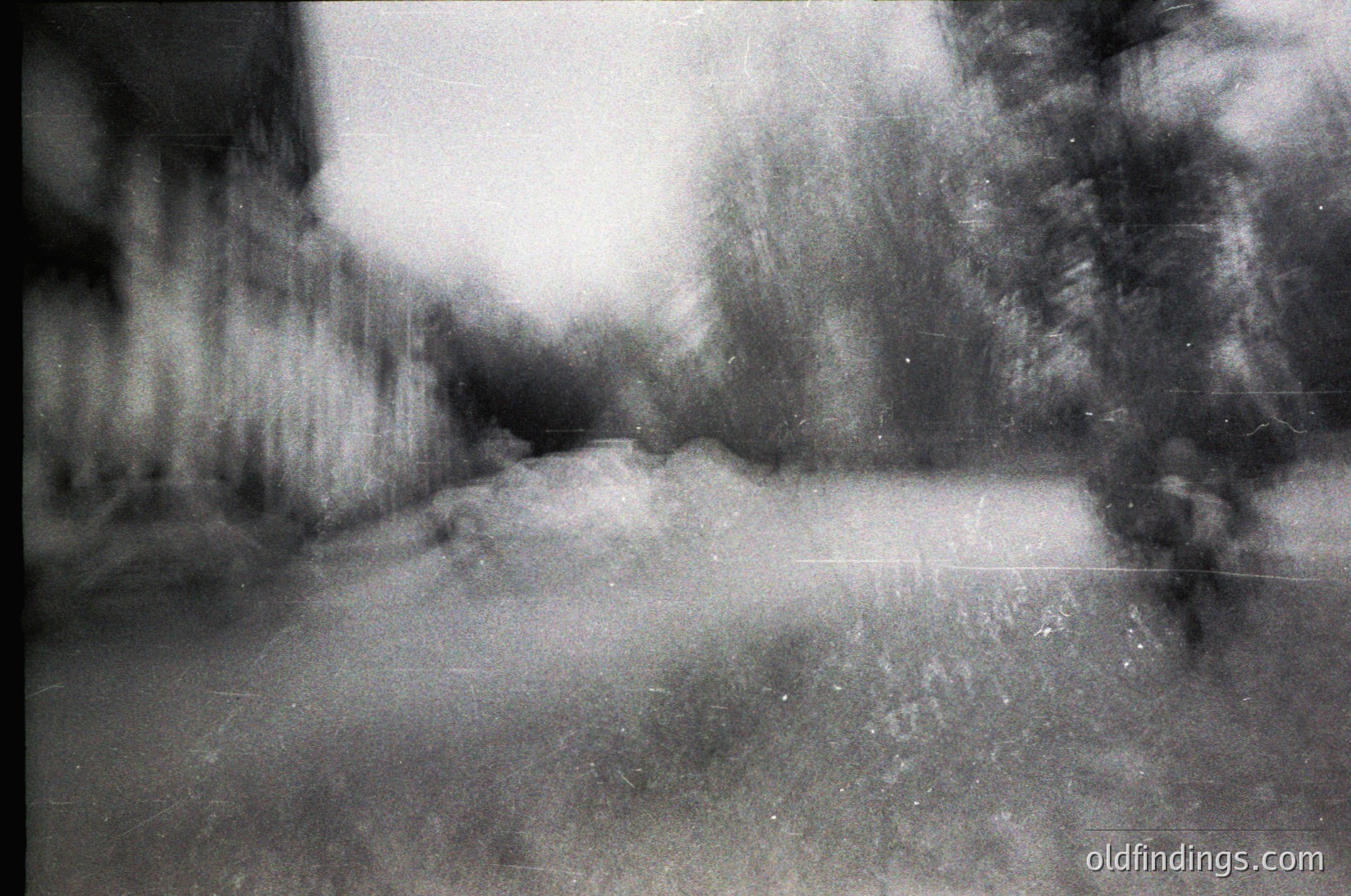 Vintage monochrome street scene with soft focus, likely 1960s–1980s. Blurred urban alleyway flanked by indistinct buildings and trees, suggesting a European or North American city. Grainy texture and faded edges enhance nostalgic, documentary-style aesthetic.