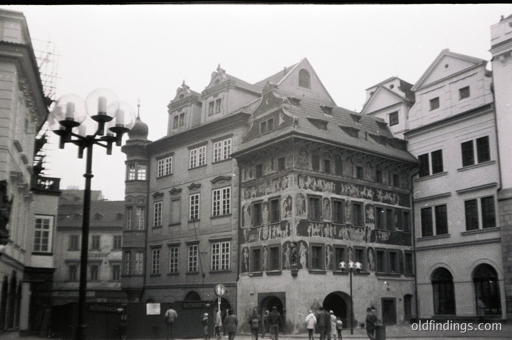 Renoir-style façade with intricate stone carvings and arched windows, likely 19th-century European urban architecture. Prominent gabled roof and decorative reliefs above the entrance. Pedestrians and vintage street lamps add context.