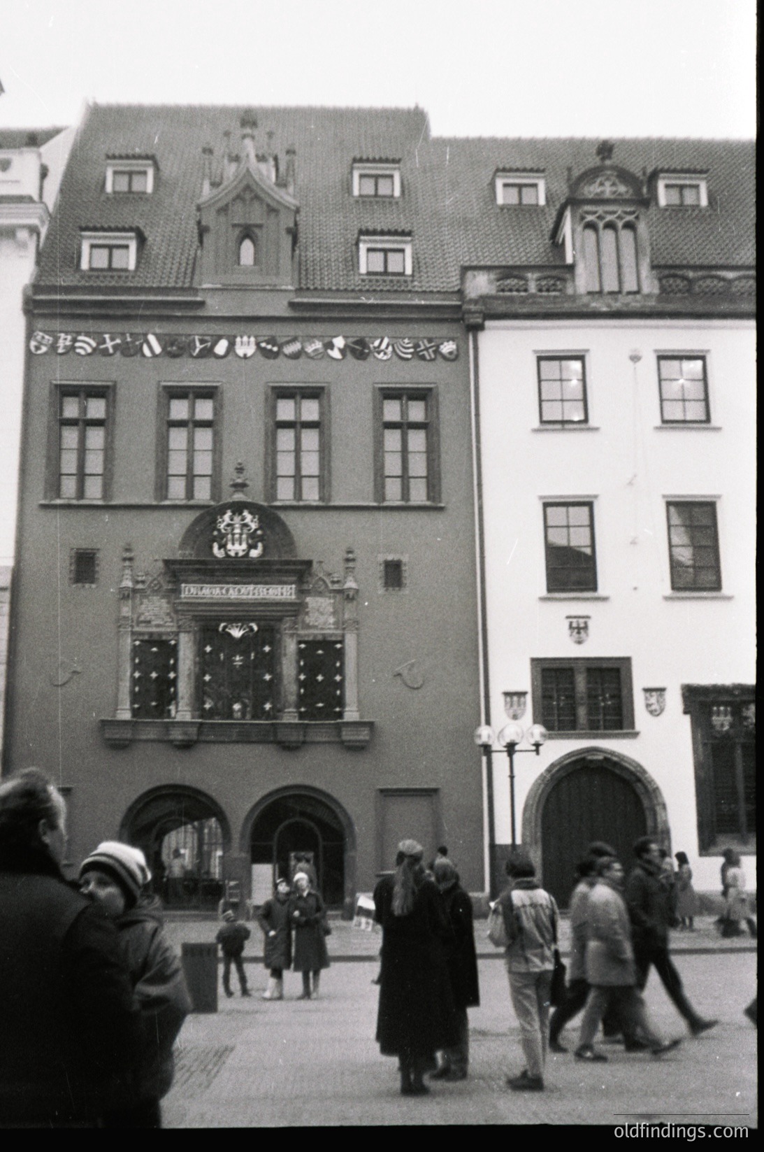 Historic European town hall façade with Gothic Revival details, including ornate crest and arched entrance. Crowd of mid-20th-century attire in a cobblestone courtyard. Likely --- *Note: Exact location indeterminable without clearer markers, but architectural style suggests Northern/Central Europe.*