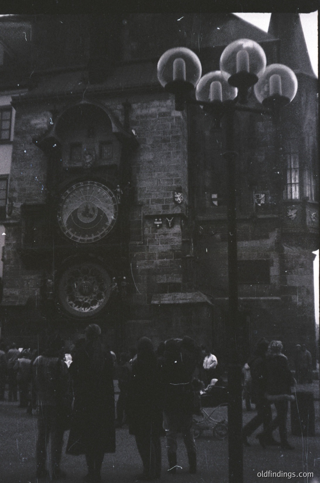 Vintage black-and-white photo of Prague’s Astronomical Clock tower (Orloj) with medieval architecture and ornate astronomical dials. Crowd of people in 1950s-era clothing gathers below ornate street lamps. Historic European urban scene.