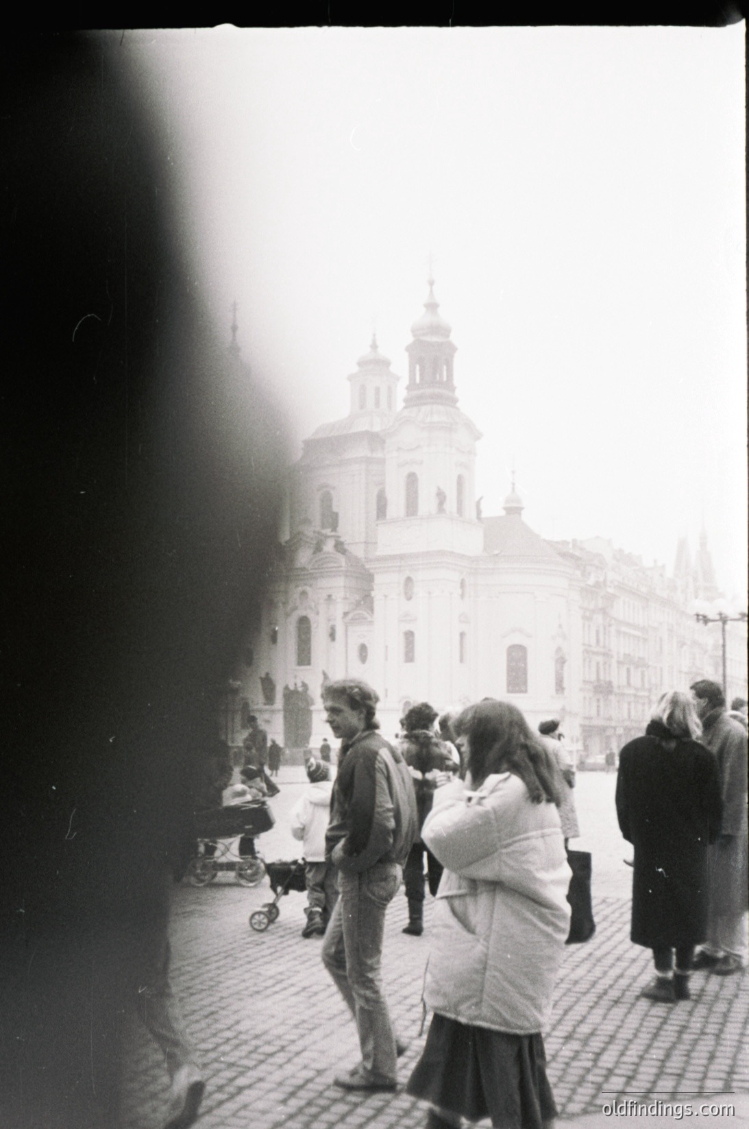 Baroque-style church with twin domes and ornate façade in a European city square, likely Prague. Crowd in 1970s–80s attire—men in jackets, women in long coats and skirts—with a stroller visible. Grainy black-and-white film captures urban life and architecture.