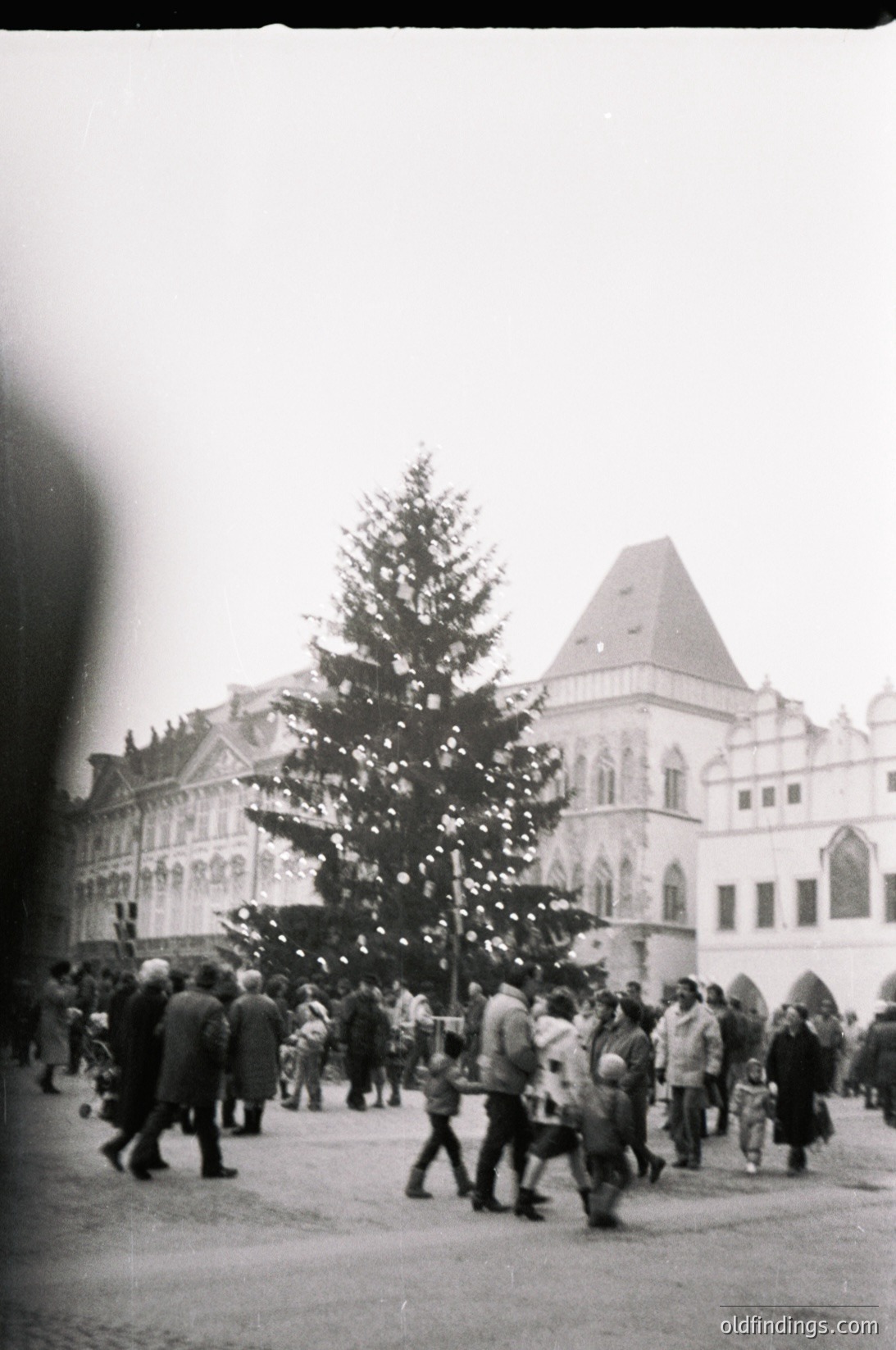 Black-and-white street scene featuring a large, decorated Christmas tree in a public square, flanked by a grand, multi-story building with ornate architecture. Crowd of people in 1960s-era clothing, including coats and hats, gathered around the tree. Urban setting with visible cobblestone ground.