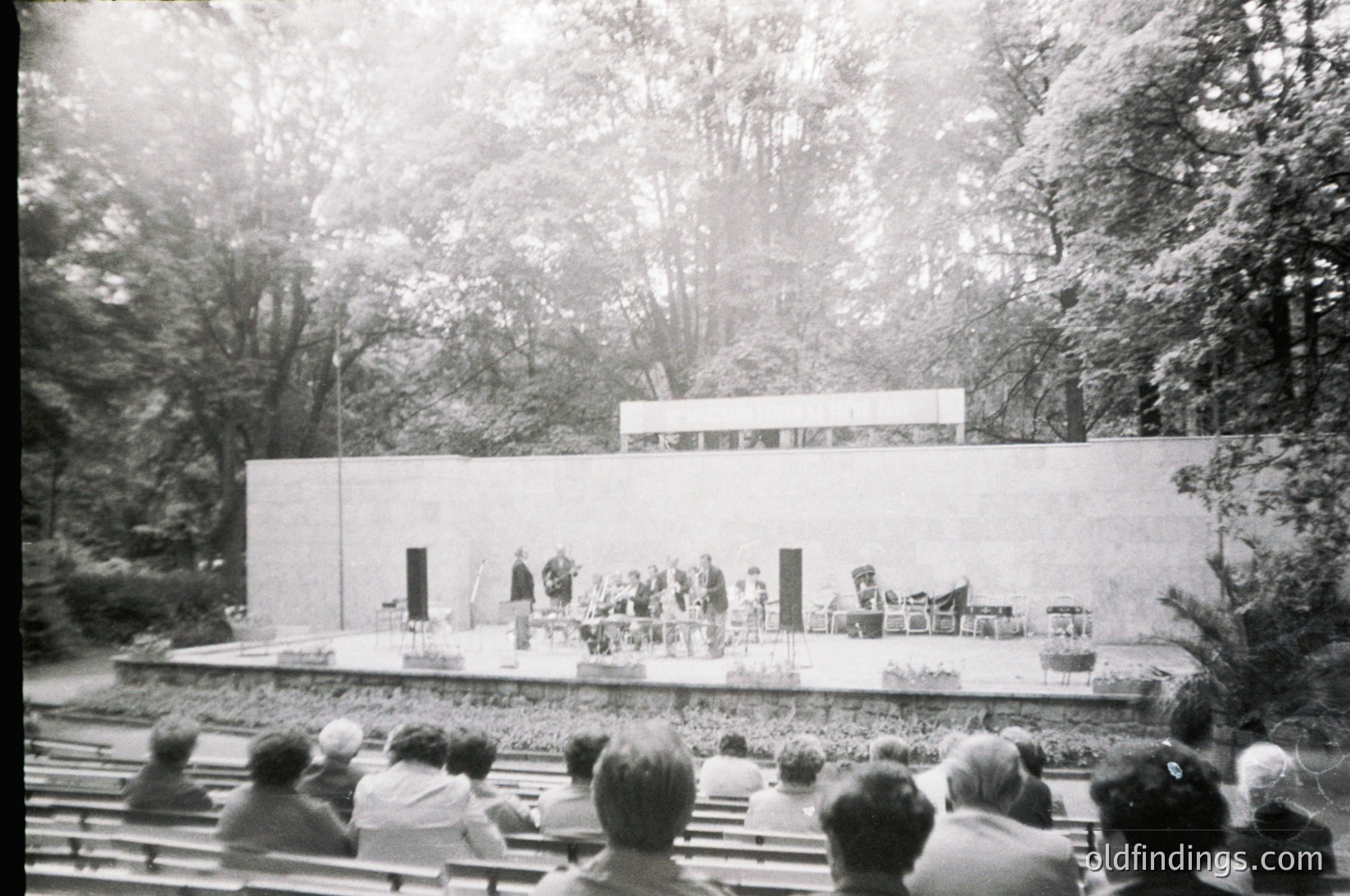 Mid-century outdoor amphitheater with tiered wooden seating under dense trees. Stage features minimalist concrete platform with two doorways and a central seating area for performers. Audience faces stage, suggesting a cultural or theatrical event. Likely 1950s–1970s based on architecture and attire.