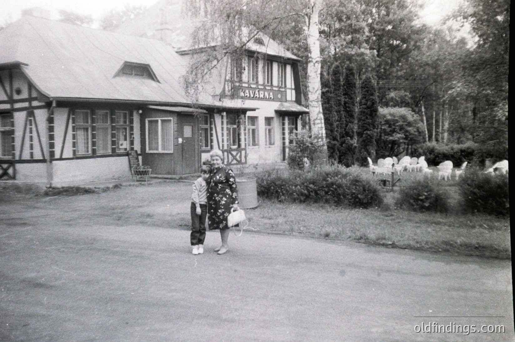 Mid-20th century black-and-white photo of a woman in traditional attire holding a basket, standing near a sign reading "Kavarna" (Bulgarian for café) in a rural or resort setting. Wooden chalet-style building with a sloped roof and large windows, surrounded by birch trees and manicured shrubs.