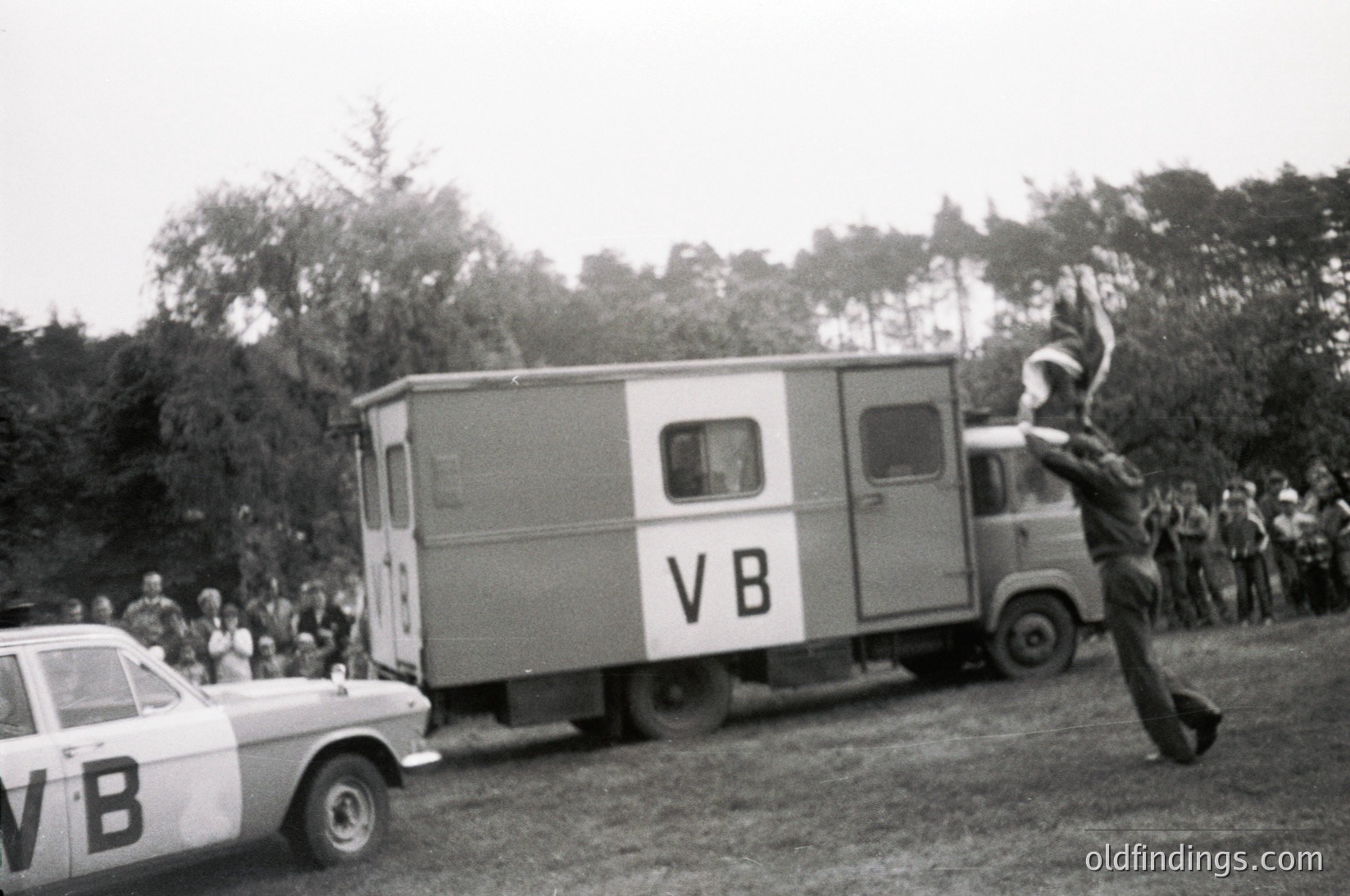 A dynamic 1960s-era photo captures a mid-air jump from a moving truck marked "VB" during a public event. Crowds gather on grassy terrain, with another "VB" vehicle parked nearby. The action suggests a promotional stunt or festival activity.