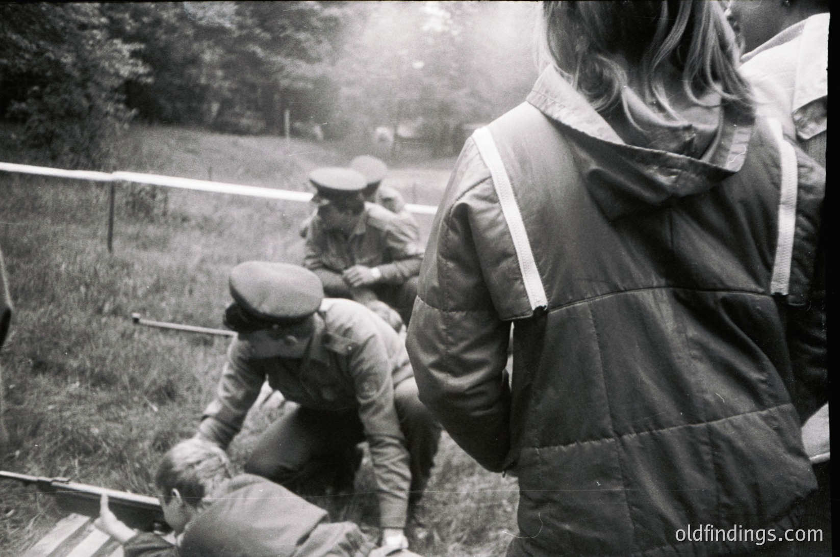 Mid-20th century outdoor scene featuring three men in military-style attire. One kneels assisting a seated individual, while another stands in the background. Fog or mist obscures distant trees, suggesting a rural or training setting. Uniforms include caps, jackets, and trousers with visible insignia.