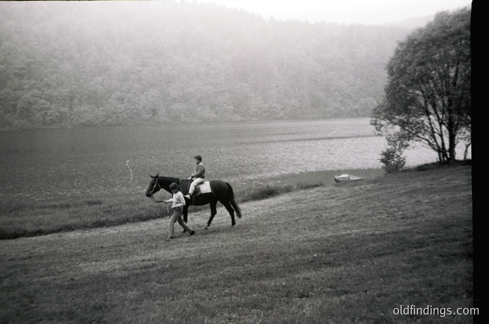 Two riders on horseback traverse a misty lakeside path, framed by dense forest. The woman leads a horse with a child seated behind, both in period attire. Likely mid-20th century, possibly , in a temperate forest region.