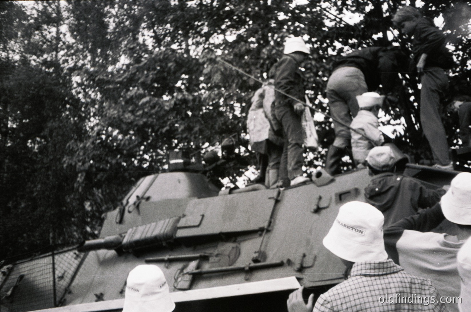 Vintage black-and-white photo of a Soviet T-34 tank with soldiers atop, likely mid-1940s Eastern Front. Crowd in white caps (possibly civilians) observes.