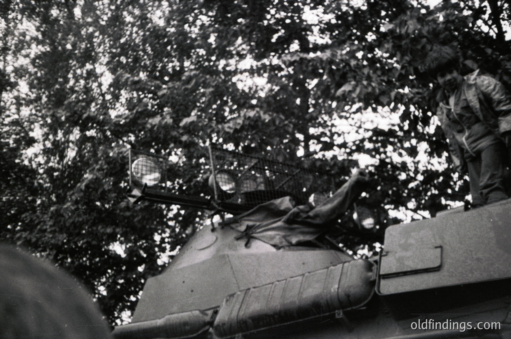 Vintage black-and-white photo of a Soviet T-34/85 tank crewman leaning over turret edge in dense forest, mid-20th century. Visible headset, periscope, and camouflage netting.
