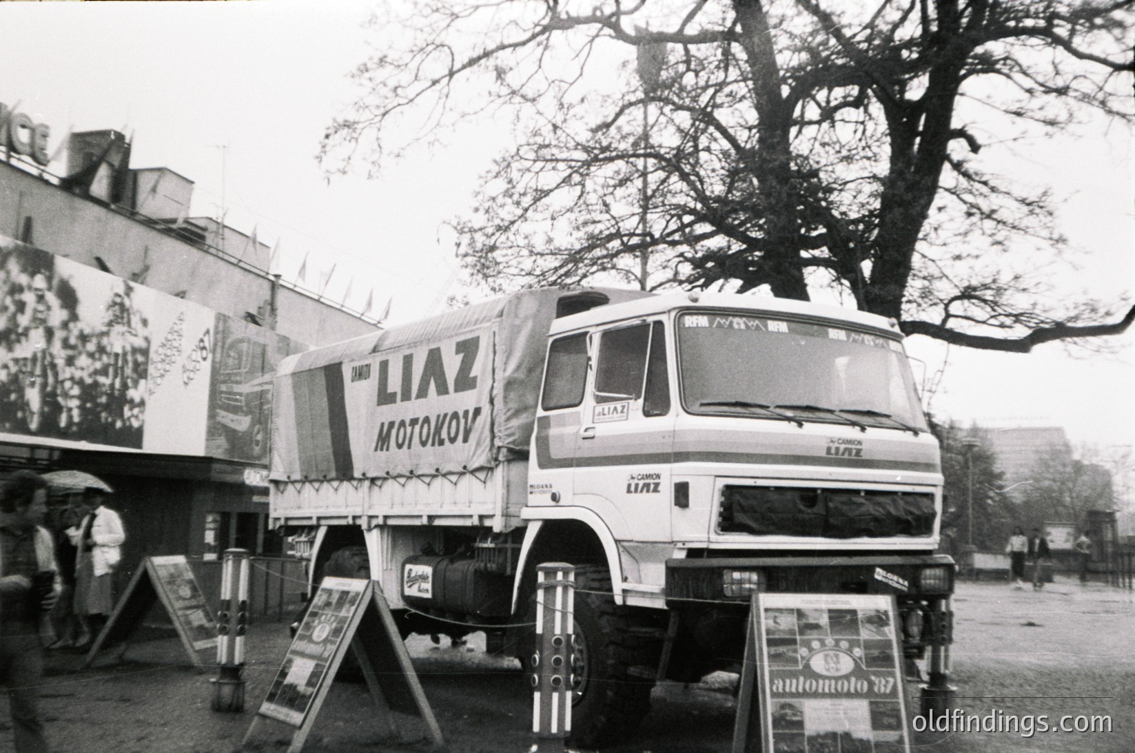 A vintage **LIAZ** truck (model 122M) from the **1970s–1980s** displays "Motokov" branding on its side, parked in an outdoor setting. The truck’s white cab and trailer feature bold Cyrillic lettering, suggesting a **Soviet/Eastern Bloc** origin. Nearby, promotional posters and a sign for "Automoto" hint at a trade show or exhibition. Urban greenery and a tree frame the scene.