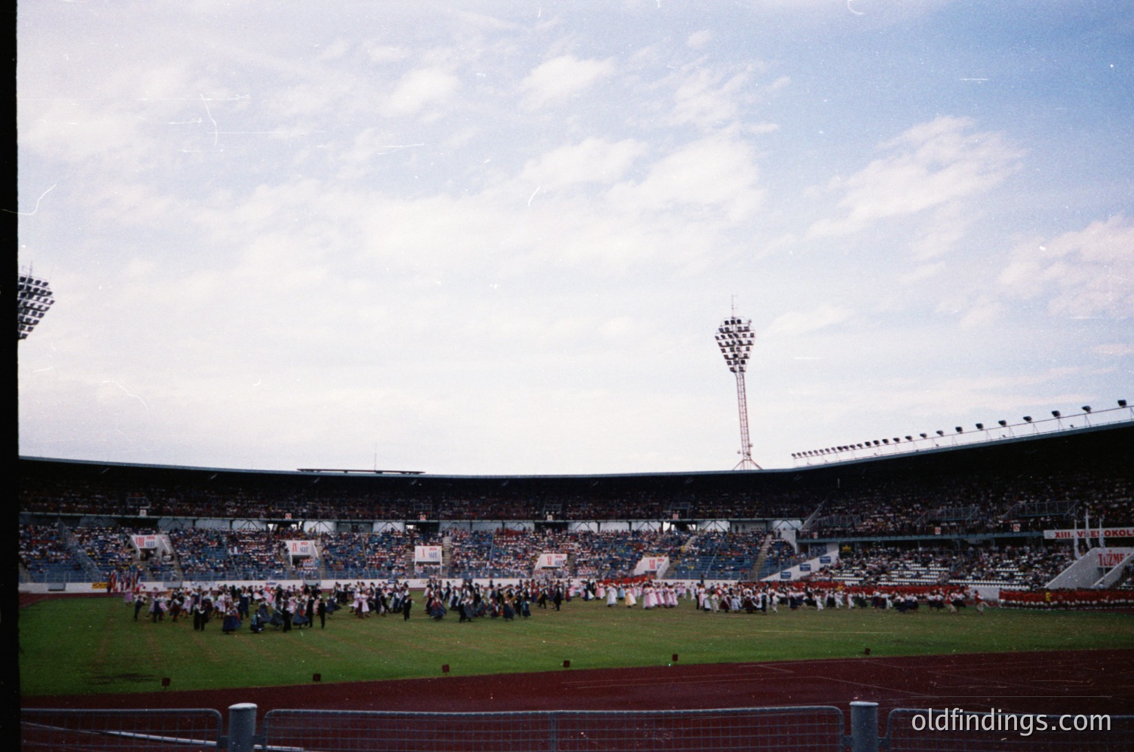Vintage stadium scene with tiered seating filled with spectators, likely a 1970s European match. Crowds hold flags and banners, suggesting a national or club event. Floodlights and a central tower frame the field.