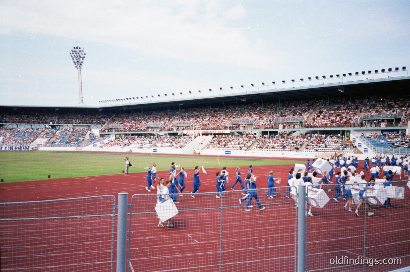 Vintage stadium scene with red artificial turf, featuring a team in blue uniforms celebrating near the sideline. Crowd fills tiered stands under a cloudy sky. Stadium architecture includes floodlights and a covered upper tier. Likely 1970s–1990s European soccer match.