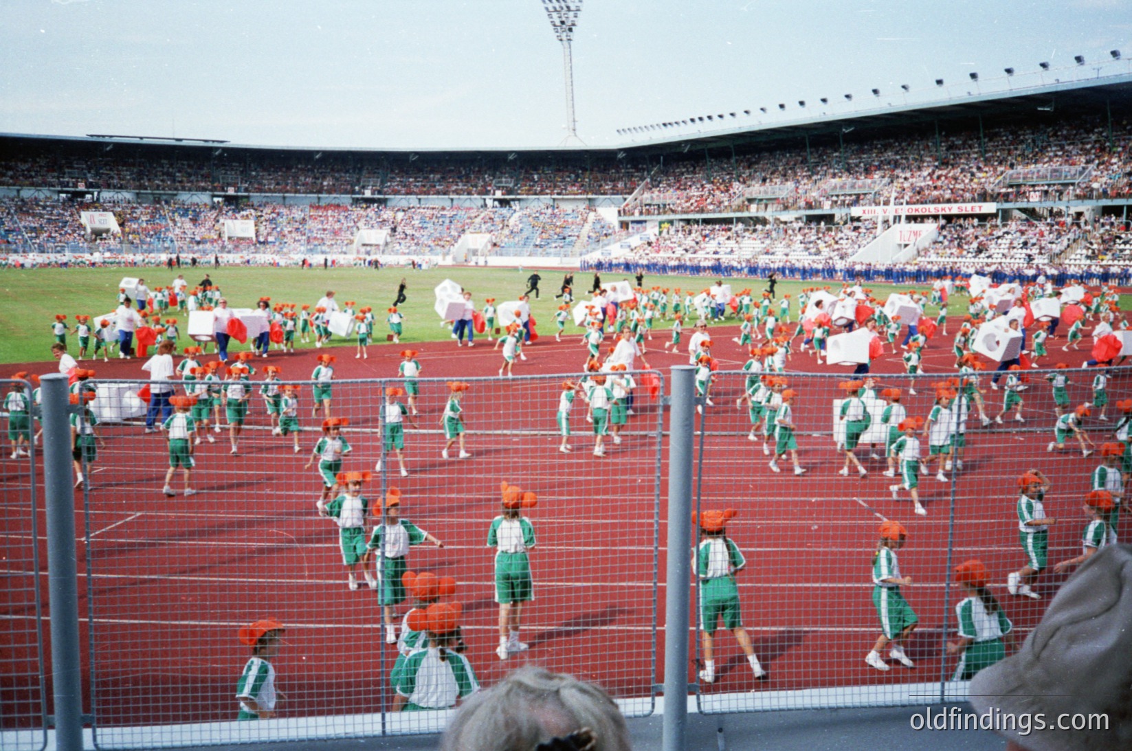Children in coordinated green-and-white sportswear, including red caps, participate in a large-scale track-and-field relay event. Stadium filled with spectators, suggesting a public/community sports day. Mid-20th century stadium design with tiered seating and floodlights.