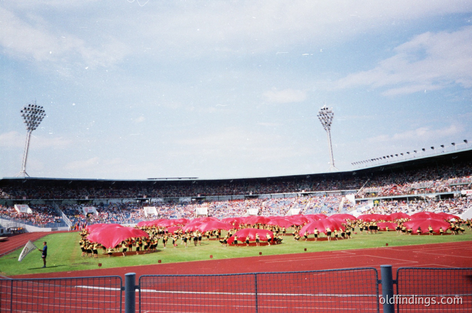 Vintage stadium scene featuring synchronized team in red uniforms and matching scarves forming a large logo on the field. Empty stands suggest pre-game or ceremonial event. Stadium lighting and floodlights indicate evening or indoor venue. Likely 1970s–1980s based on attire and lighting.