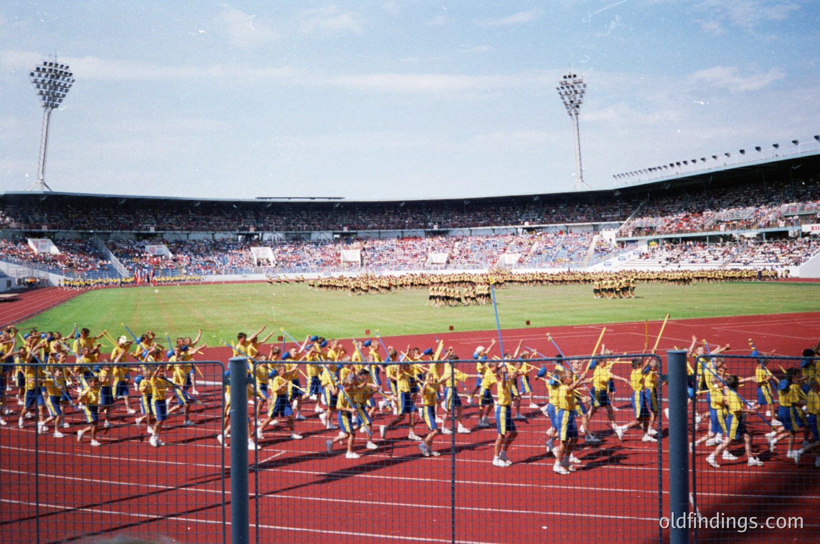 A large-scale synchronized marching band performance in a stadium, featuring uniformed musicians in yellow jackets and blue pants. The event appears to be part of a major sports or cultural celebration, with a packed crowd in the stands holding flags. The stadium’s red track and floodlights suggest an evening or early afternoon setting. Likely a 1970s–1980s European sports event, possibly a World Cup or Olympic opening ceremony.