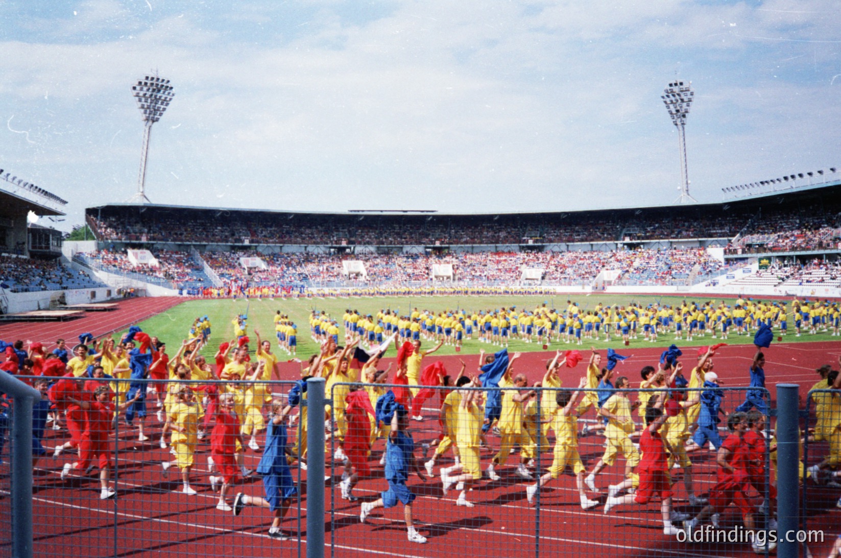 Massive stadium scene with synchronized marching band in vibrant red/yellow/blue uniforms performing on track. Crowd fills stands, suggesting major event. Stadium lighting and floodlights indicate evening or indoor venue. Likely 1970s–1980s based on uniforms and stadium design.