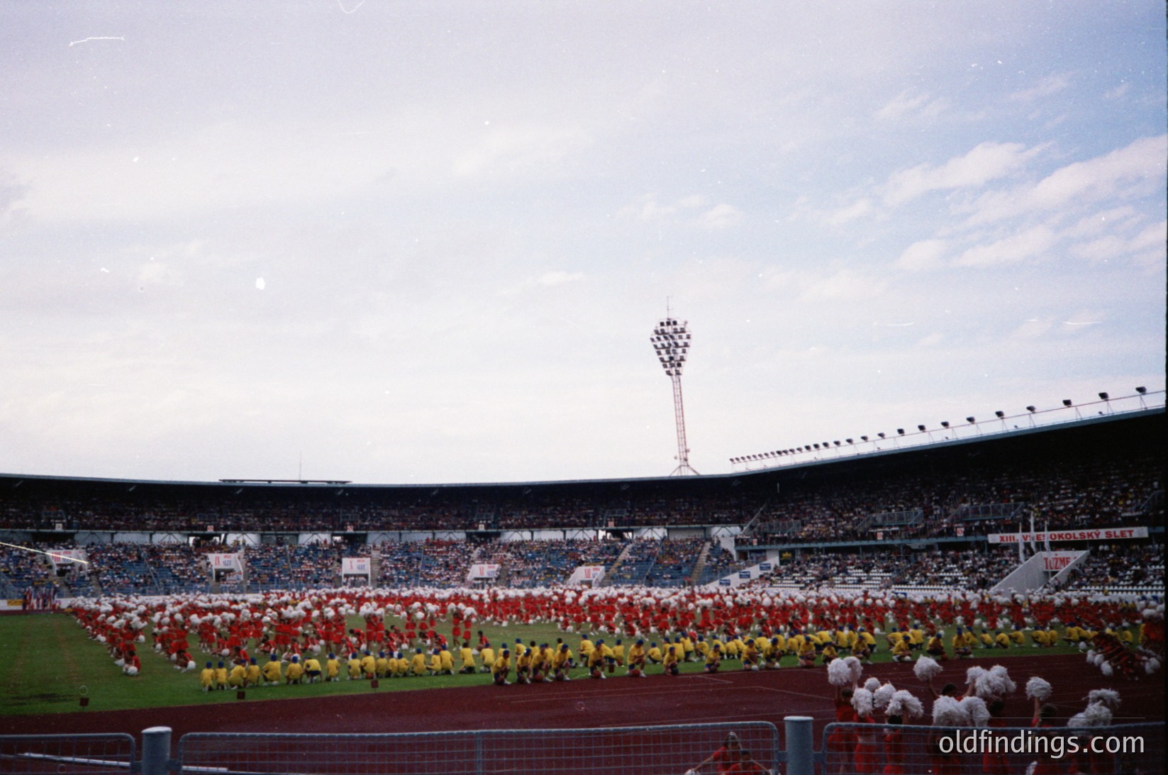 Large stadium ceremony with coordinated teams in red, yellow, and white uniforms forming geometric patterns on the field. Crowds fill tiered seating under a tall scoreboard. Mid-20th century European sports venue, likely or .