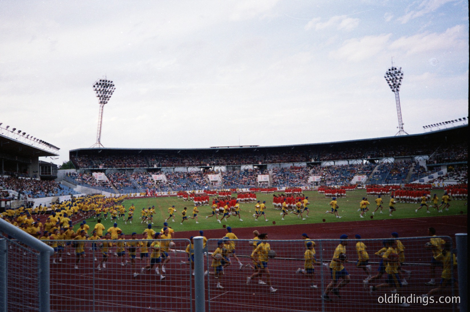 Vintage stadium scene featuring synchronized team drills in yellow jerseys on grass field. Crowd in stands, partially obscured by fencing. Stadium lighting and floodlights visible. Likely 1970s-1980s European football culture.