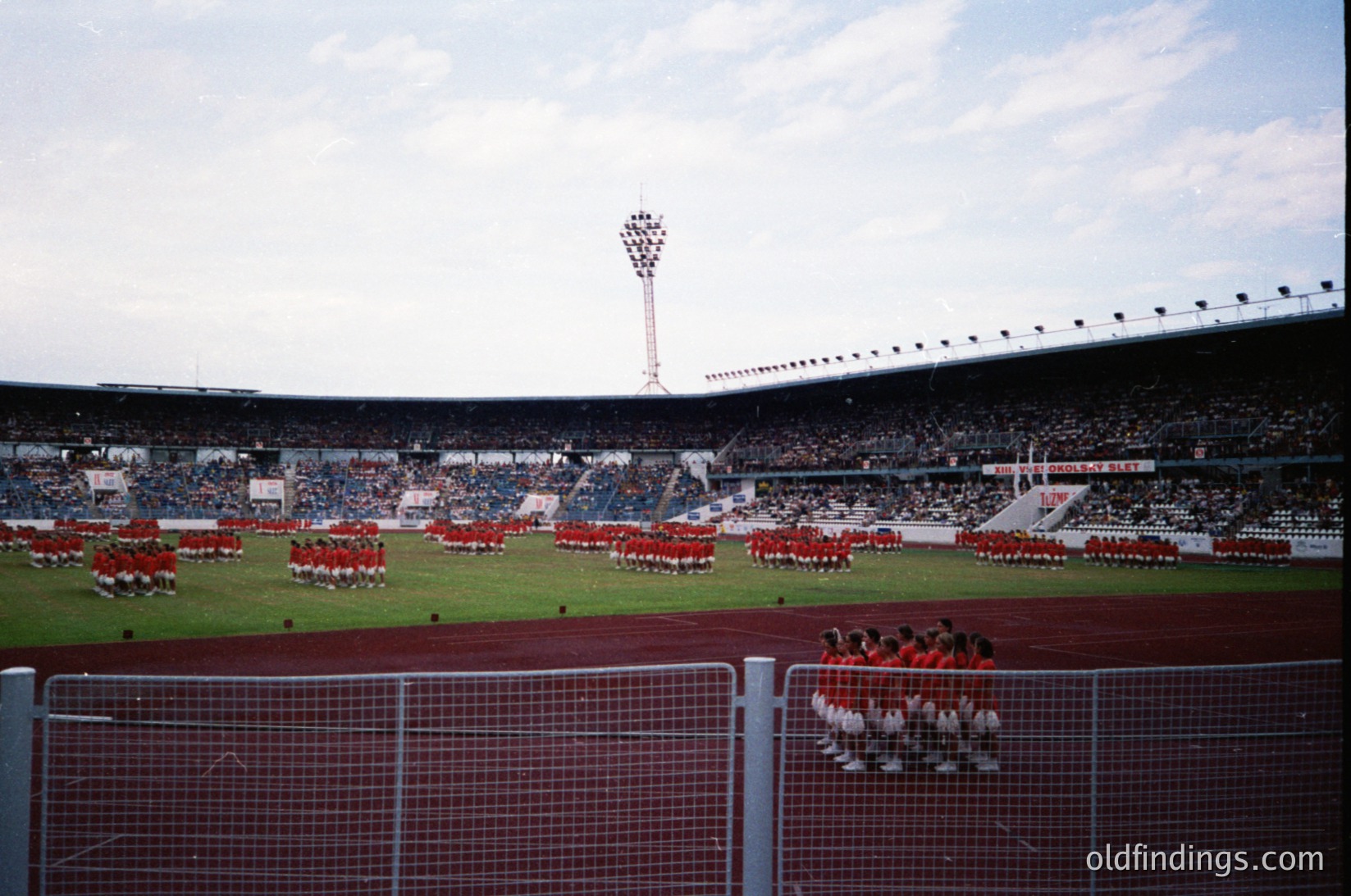 Massive stadium event featuring synchronized marching bands in coordinated red/white uniforms. Crowds fill tiered seating under a tall floodlight tower. Open-air venue with clear skies, likely a sports or cultural festival. Mid-20th century stadium architecture with high seating capacity.