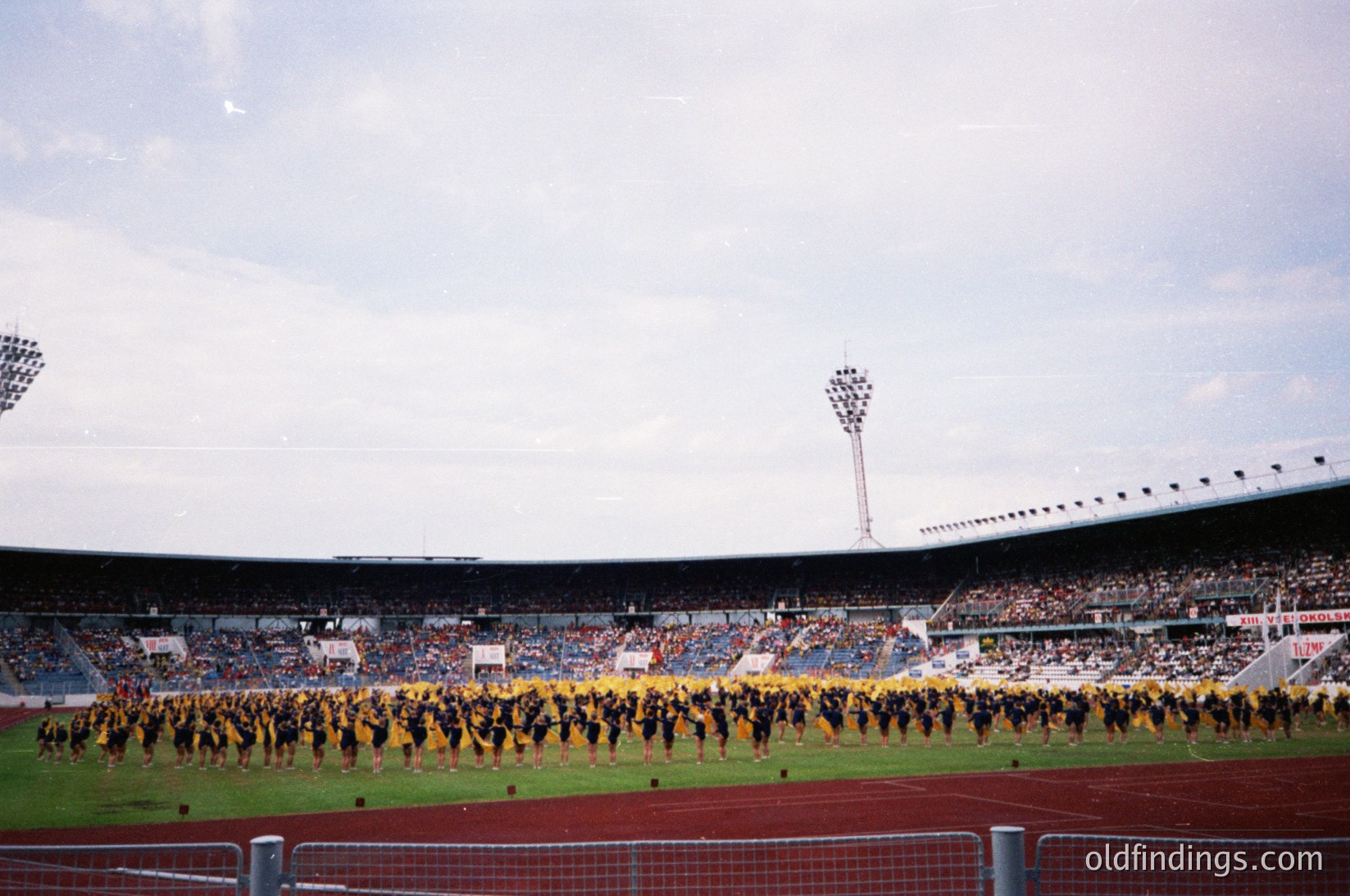 Massive stadium crowd with coordinated cheerleaders in yellow and black performing on field. Stadium seating filled with spectators under cloudy skies. Likely 1970s–1990s European sports event.