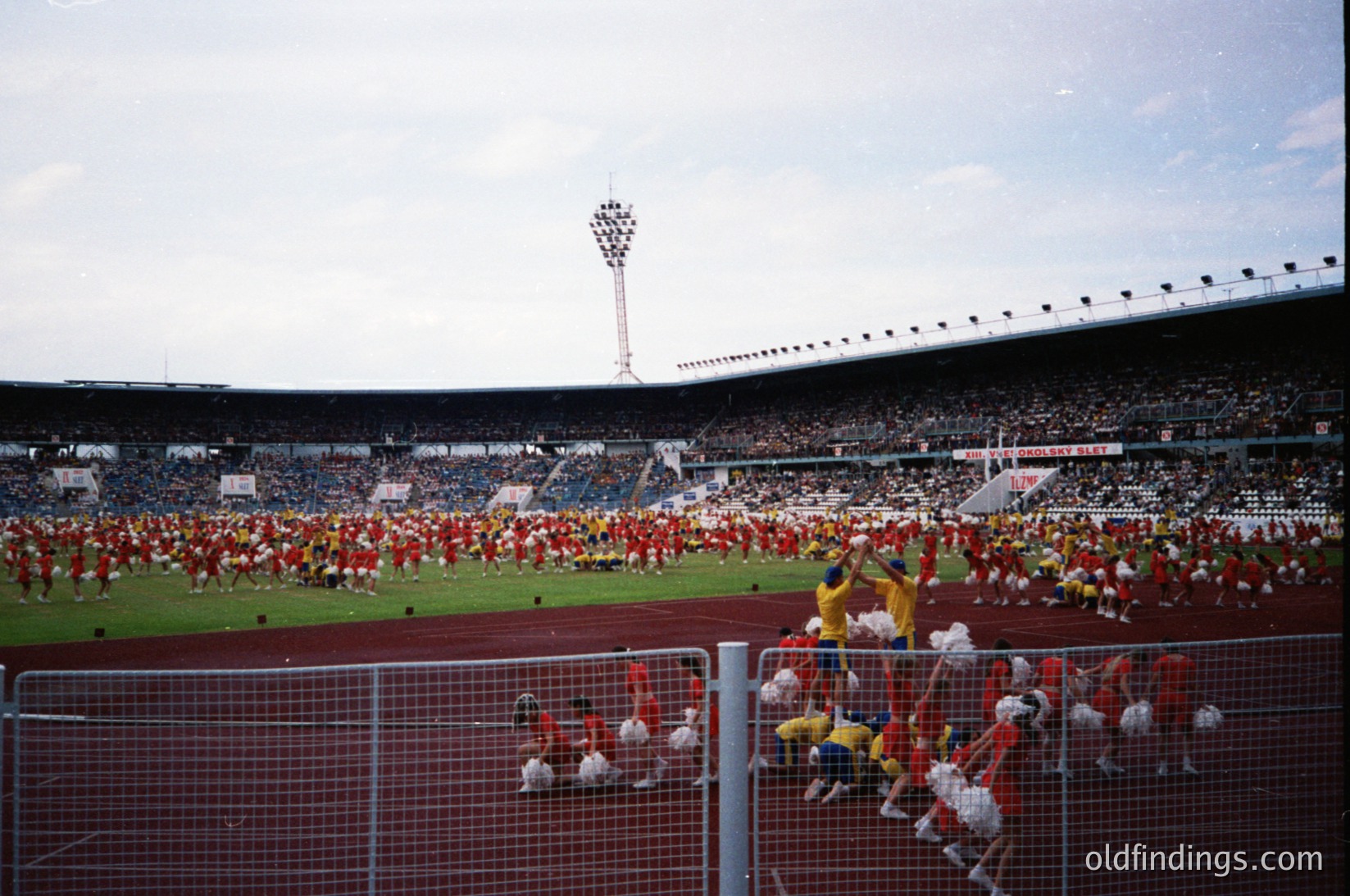 Massive stadium scene featuring synchronized cheerleaders in red and white uniforms performing on the field. Crowd fills tiered seating under stadium lights. Likely 1970s–1980s sports event, possibly European.