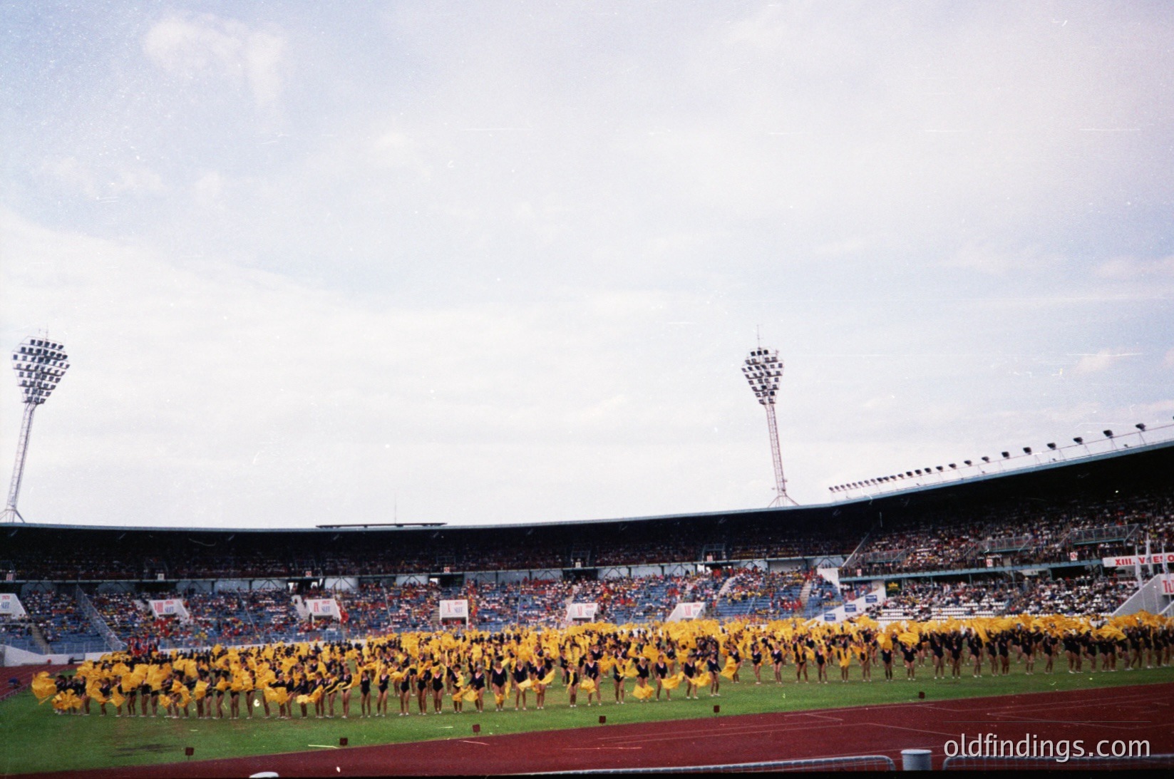 Vintage stadium scene featuring a large synchronized marching band in yellow uniforms performing on a grass field. Stadium seating partially filled with spectators under floodlights. Likely a 1960s–1970s European sports event.