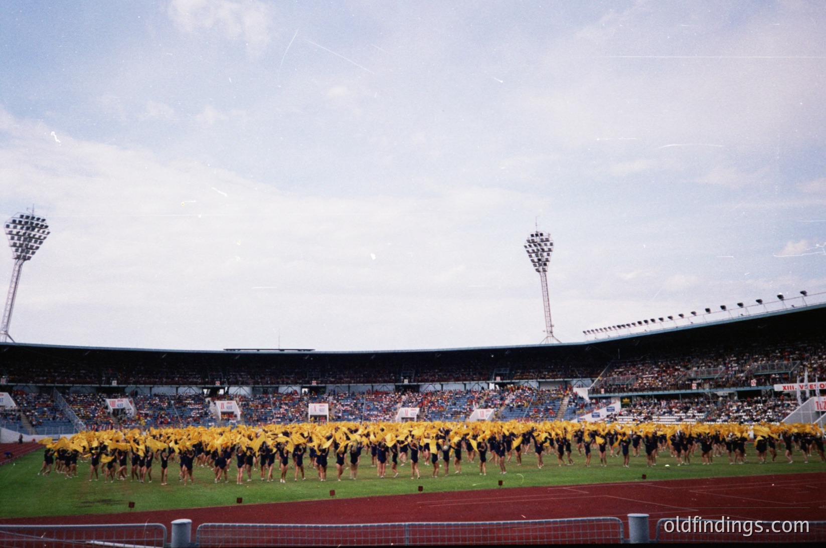 Vintage stadium scene featuring a synchronized dance routine by uniformed performers in yellow and black. Empty stands contrast with the vibrant formation on the field. Classic floodlights and tiered seating suggest a mid-20th-century European venue.