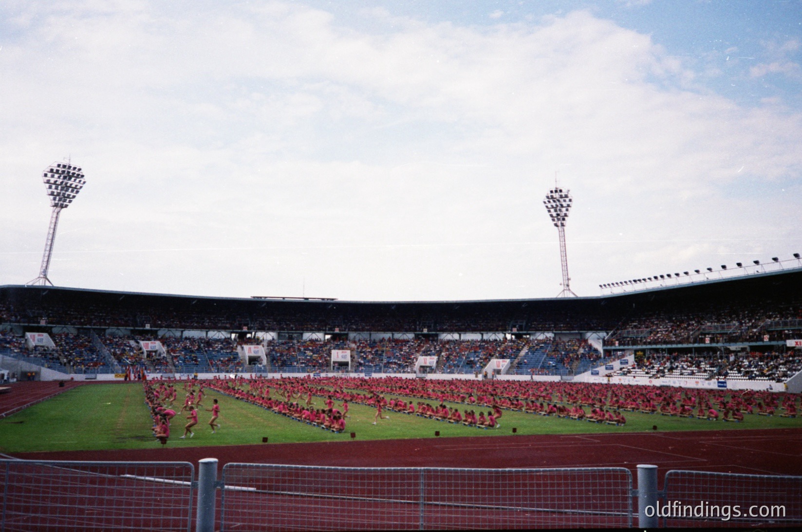 Large stadium crowd performing synchronized choreography in red uniforms, likely a sports or ceremonial event. Mid-20th century stadium design with floodlights and tiered seating. Empty field area in foreground suggests pre-game or halftime display.
